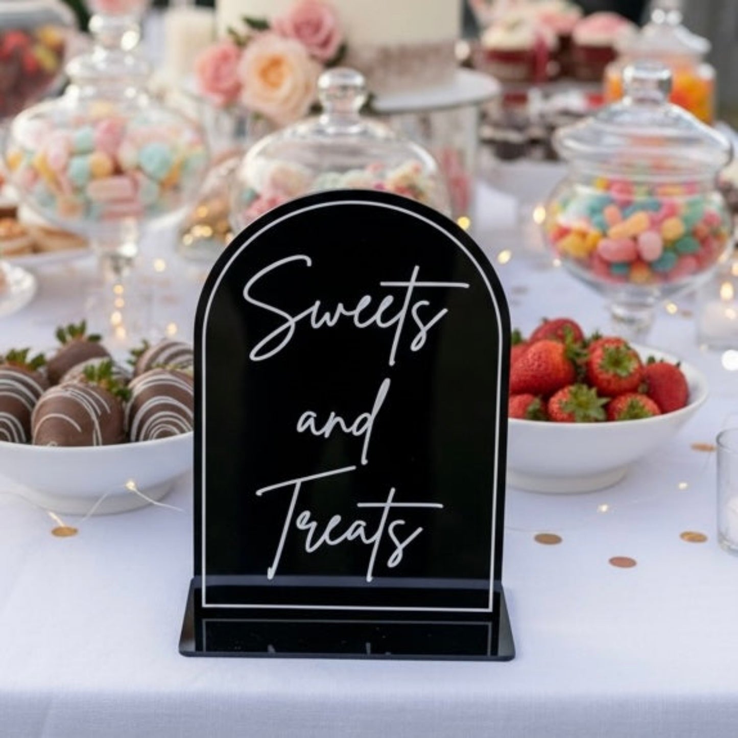 A black acrylic sign with white script reading 'Sweets and Treats' standing on a white tablecloth at a dessert table with bowls of strawberries, chocolate-covered treats, and candy under glass domes.