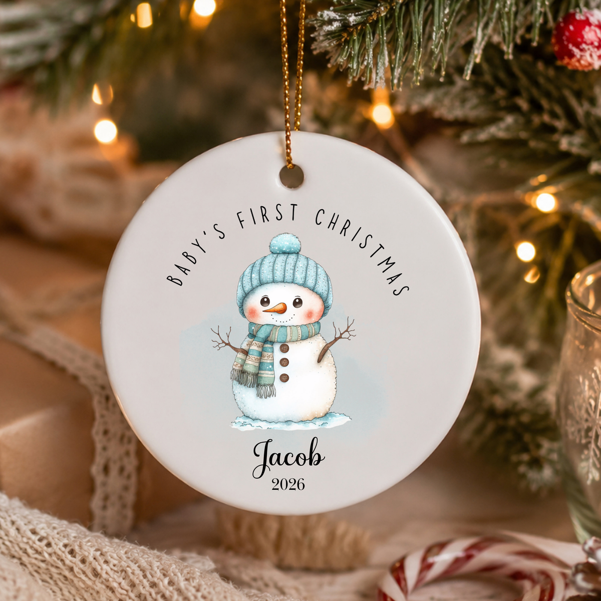 A round white ceramic Christmas ornament hanging from a gold string, featuring a watercolor snowman wearing a blue knit hat and scarf. The ornament is personalized with the text 'BABY'S FIRST CHRISTMAS' and the name 'Jacob 2026'.