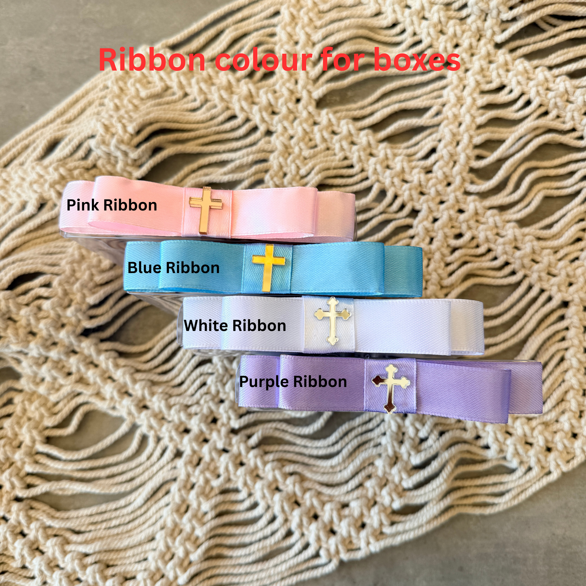 Four satin ribbon boxes in pink, blue, white, and purple, each with a small gold cross charm, arranged on a beige woven textile. Red text at the top reads 'Ribbon colour for boxes'.