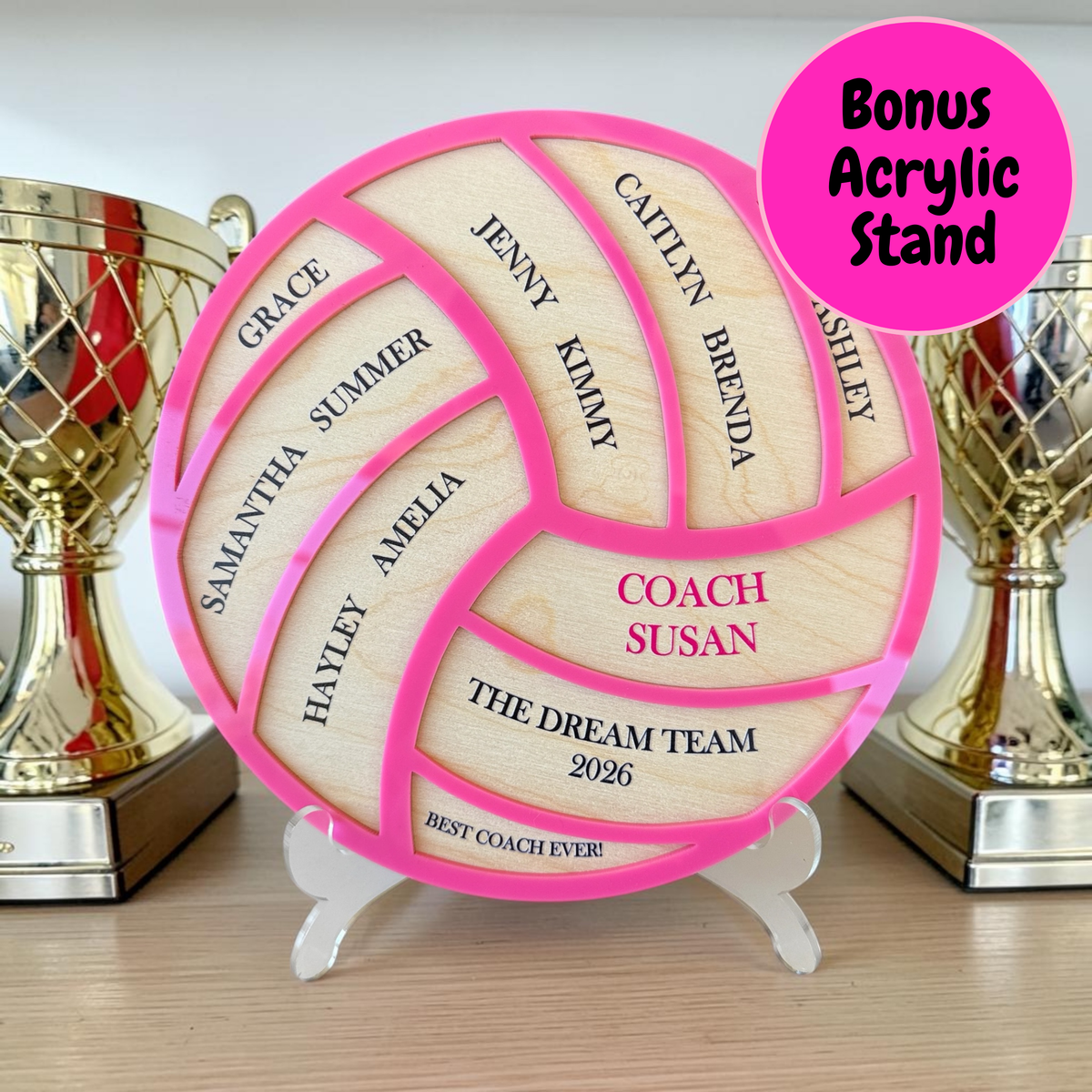 Personalized wooden volleyball-shaped plaque on a clear acrylic stand, with bright pink sections and engraved names and text including 'COACH SUSAN' and 'THE DREAM TEAM 2026'. Two gold trophies sit in the background on a tabletop, and a pink circular badge in the top right says 'Bonus Acrylic Stand'.