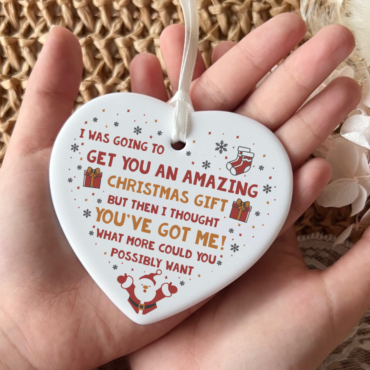 A heart-shaped white ceramic Christmas ornament held in a hand, featuring a humorous printed message in red and orange text with small red and green holiday illustrations, including a Santa Claus, stocking, and gift boxes.