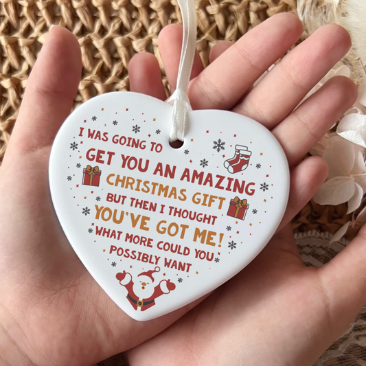 A heart-shaped white ceramic Christmas ornament held in a hand, featuring a humorous printed message in red and orange text with small red and green holiday illustrations, including a Santa Claus, stocking, and gift boxes.