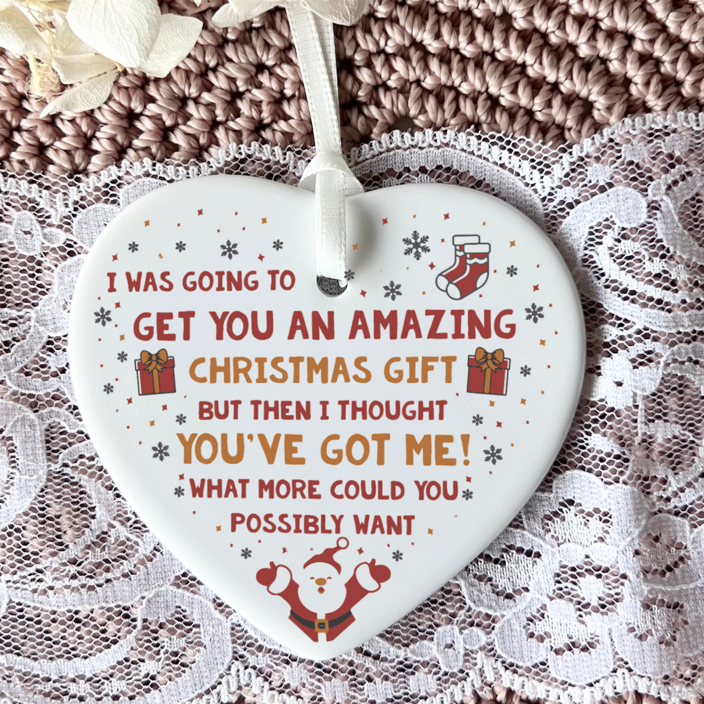 A heart-shaped white ceramic Christmas ornament with humorous text, featuring red and orange lettering, a Santa graphic, and small gift and stocking illustrations, hanging against a textured brown knit and lace background.