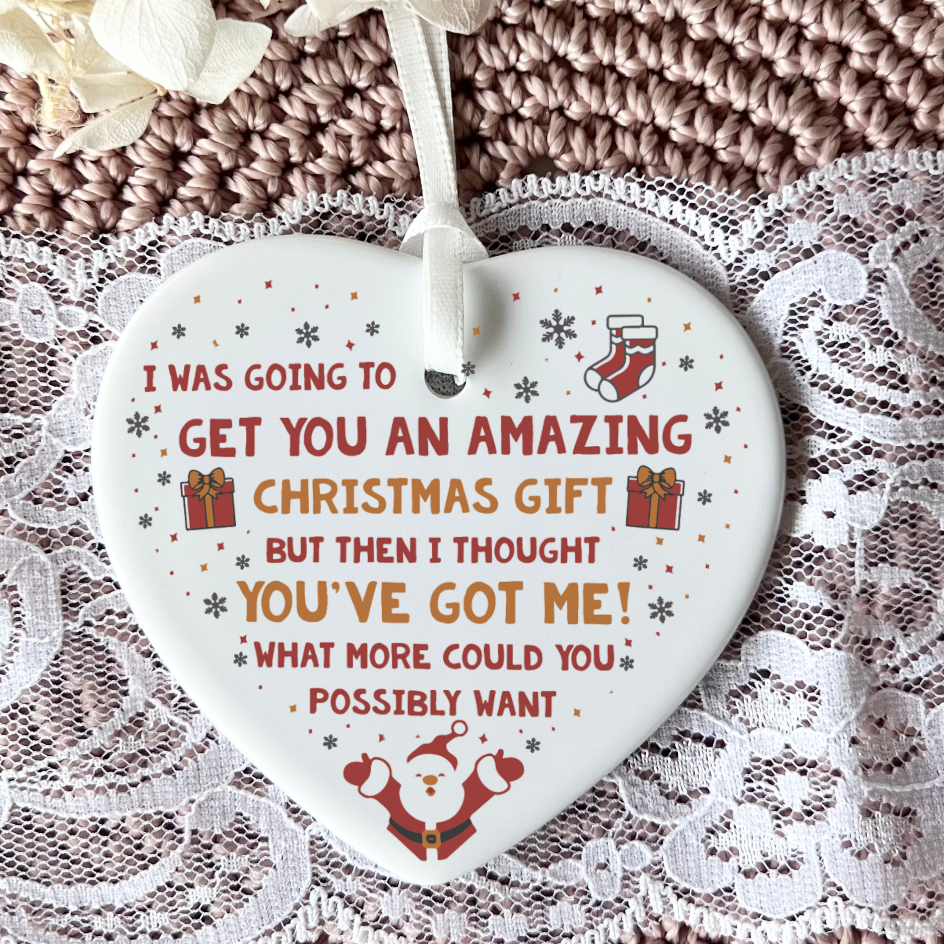 A heart-shaped white ceramic Christmas ornament with humorous text, featuring red and orange lettering, a Santa graphic, and small gift and stocking illustrations, hanging against a textured brown knit and lace background.