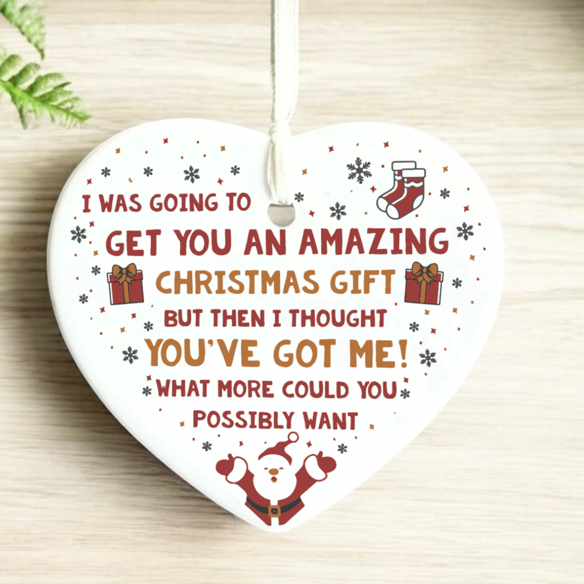 A heart-shaped white ceramic Christmas ornament with a hanging loop, printed with a humorous quote in red and brown lettering, decorated with small gift boxes, a stocking, and Santa at the bottom, shown on a light wood surface.