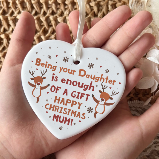 A white heart-shaped ceramic Christmas ornament held in a hand, featuring the text 'Being your Daughter is enough OF A GIFT HAPPY CHRISTMAS MUM!' in orange, decorated with two cartoon reindeer and small snowflakes.