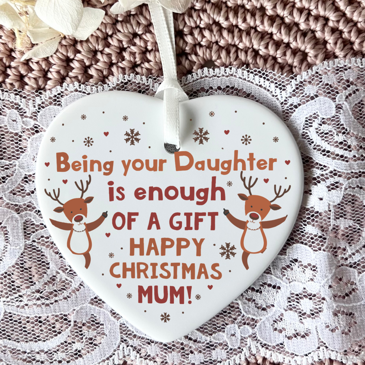 A heart-shaped ceramic Christmas ornament with the text 'Being your Daughter is enough OF A GIFT HAPPY CHRISTMAS MUM!' in red and brown lettering, decorated with two cartoon reindeer and snowflakes on a white background. The ornament is hung by a white ribbon and displayed on a lace cloth over a brown knitted blanket.