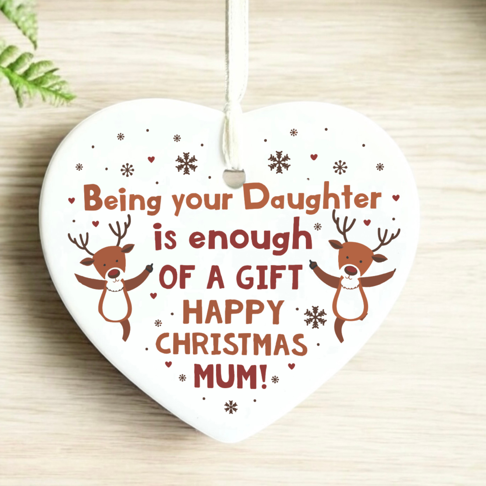 A heart-shaped white ceramic Christmas ornament hanging from a white ribbon, featuring the text 'Being your Daughter is enough OF A GIFT HAPPY CHRISTMAS MUM!' in brown and red lettering, decorated with two cartoon reindeer and snowflakes, set against a light wood background.