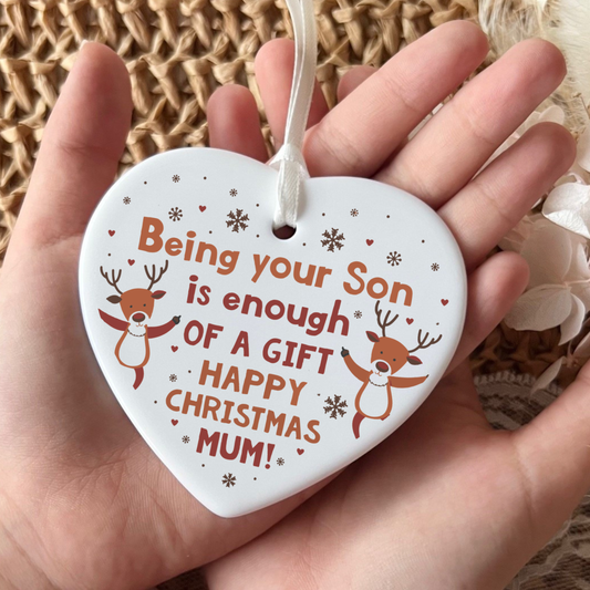 A white heart-shaped ceramic Christmas ornament held in a hand, featuring the text 'Being your Son is enough OF A GIFT HAPPY CHRISTMAS MUM!' in orange lettering, decorated with two cartoon reindeer and small snowflakes and hearts.