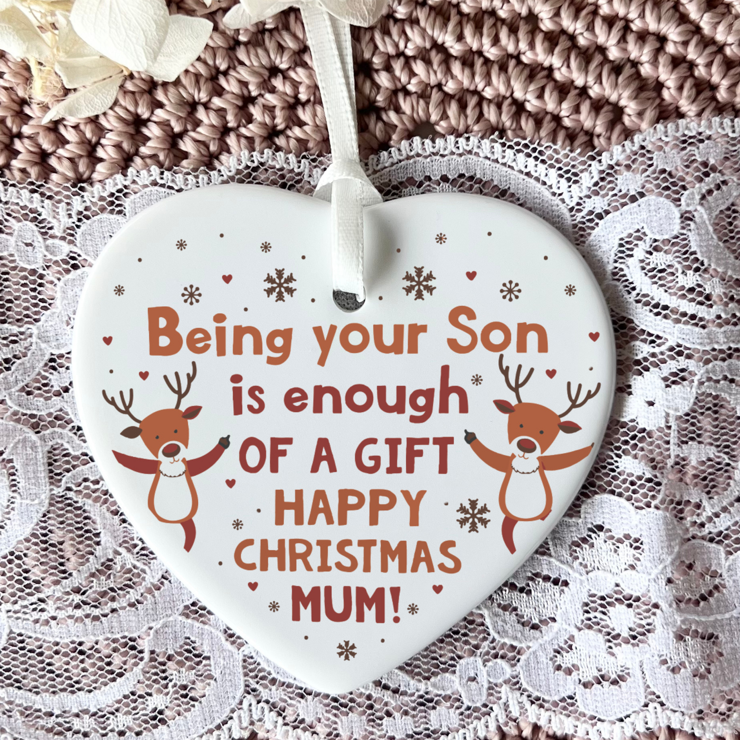 A heart-shaped white ceramic Christmas ornament with the message 'Being your Son is enough OF A GIFT HAPPY CHRISTMAS MUM!' in orange lettering, decorated with cartoon reindeer and snowflakes, hanging from a white ribbon against a textured brown knit and lace background.