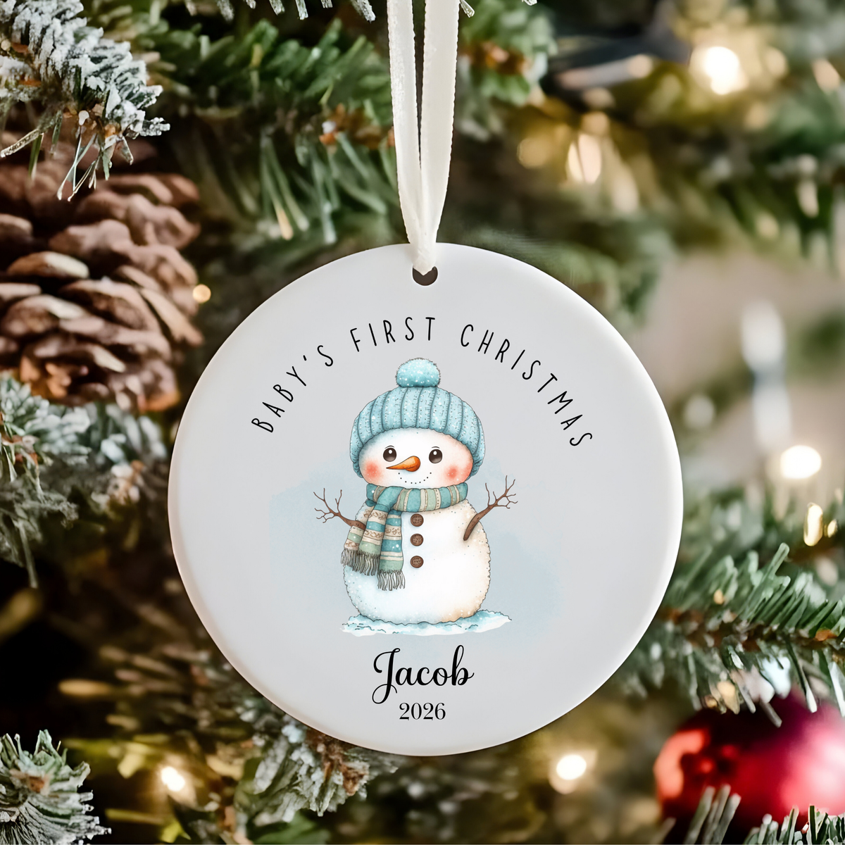 A personalized round ceramic Christmas ornament hanging on a decorated tree. The white ornament features a printed snowman wearing a blue knit hat and scarf, with the text 'BABY'S FIRST CHRISTMAS' above and 'Jacob 2026' below.