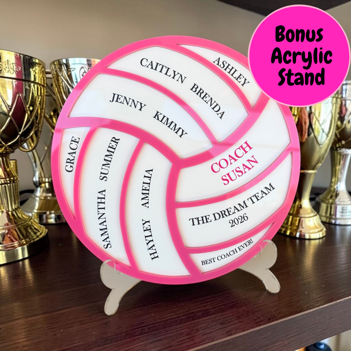 A personalized round acrylic football-shaped plaque on a small stand, displayed on a wooden shelf in front of trophies. The plaque has a bright pink border and white panels with custom names and text, including 'COACH SUSAN' and 'THE DREAM TEAM 2026'.
