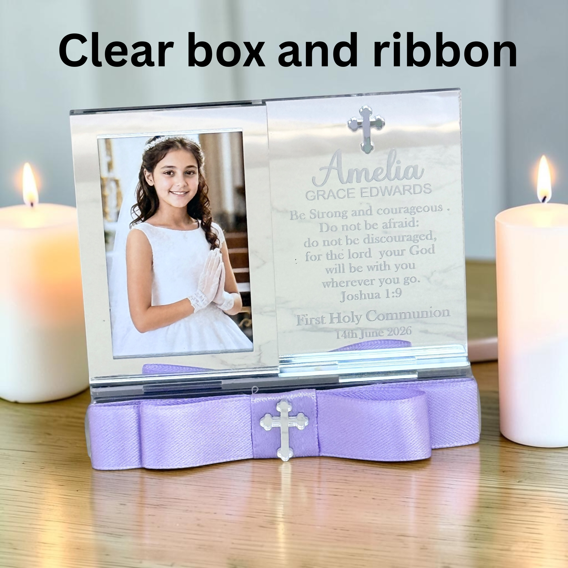 A clear box and lavender ribbon First Holy Communion photo frame stand on a wooden table, flanked by two lit white candles. The clear front panel displays a photo of a young girl in a white communion dress and includes engraved text and a small cross.