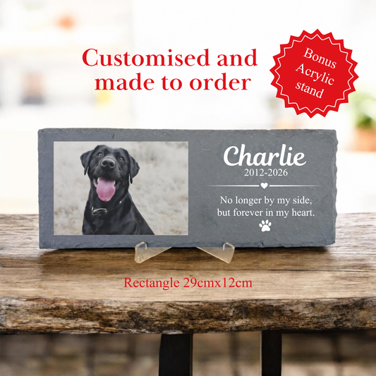 A personalized dark gray slate pet memorial plaque on a small clear acrylic stand, placed on a rustic wooden table. The plaque features a photo of a black dog and the custom text 'Charlie 2012-2026 No longer by my side, but forever in my heart.