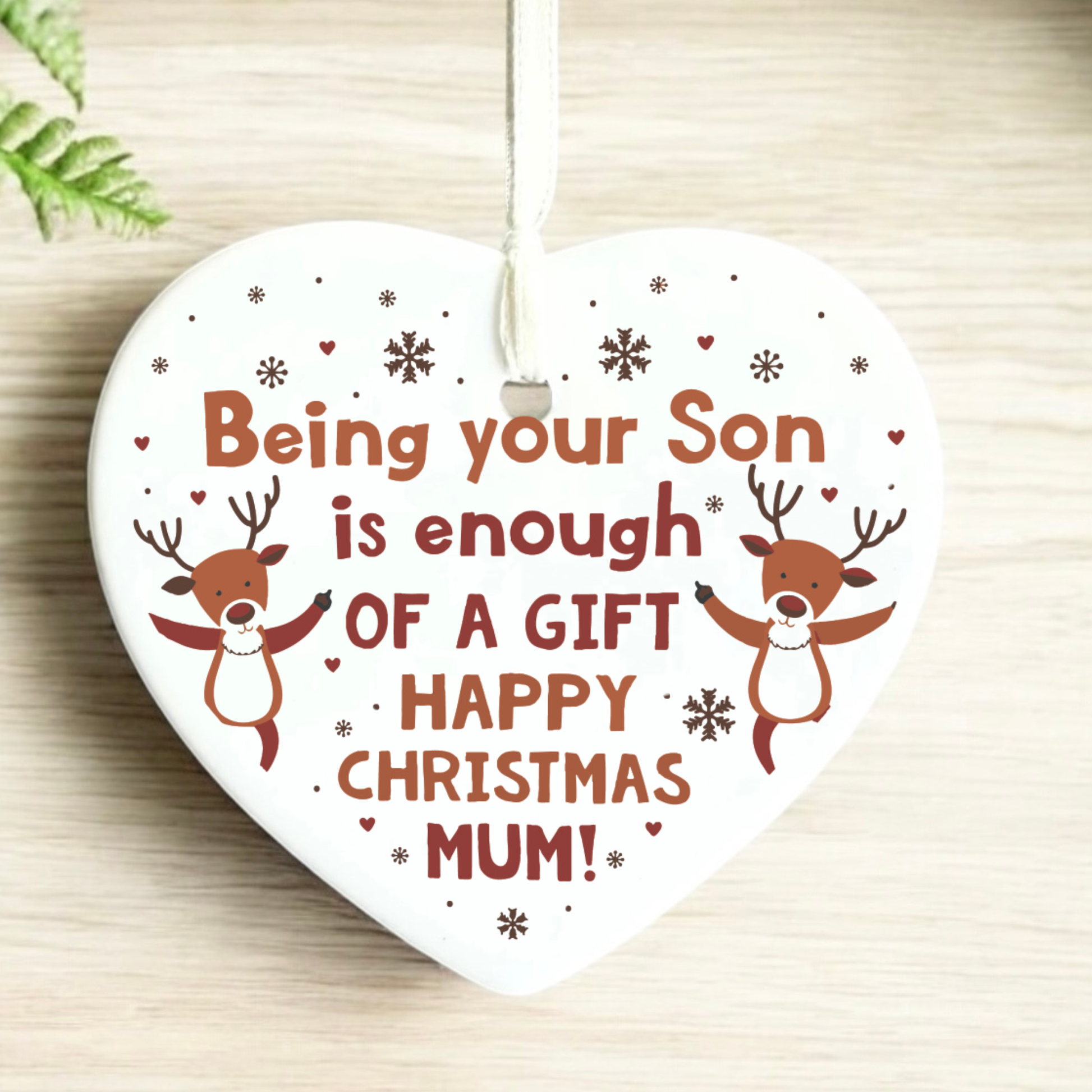 A heart-shaped white Christmas ornament with a hanging loop, featuring the text 'Being your Son is enough OF A GIFT HAPPY CHRISTMAS MUM!' in brown and red lettering, decorated with two cartoon reindeer and small snowflakes and hearts, shown on a light wood surface.