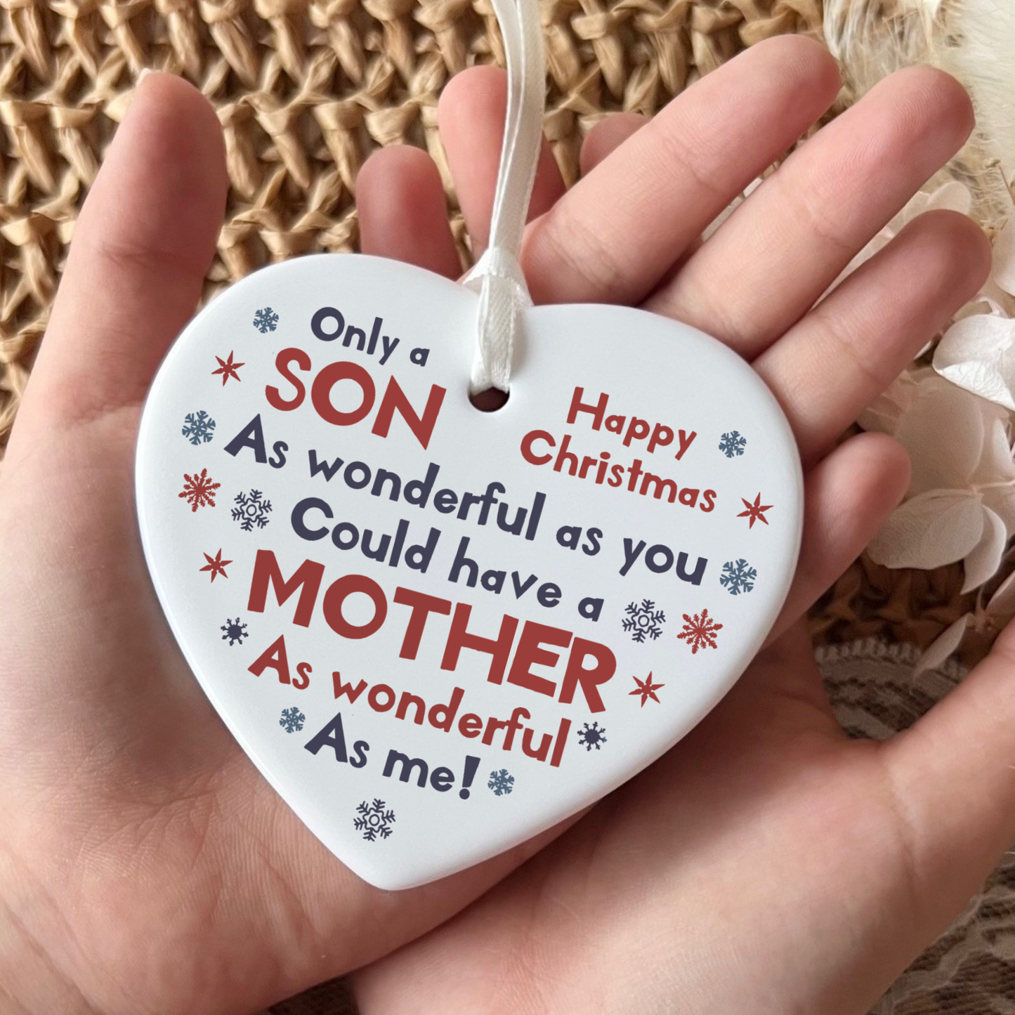 White ceramic heart-shaped Christmas ornament held in a hand as shown in 31.jpg. The ornament features red and blue snowflakes with the printed text: Only a SON As wonderful as you Could have a MOTHER As wonderful As me! Happy Christmas. It hangs from a delicate white ribbon.