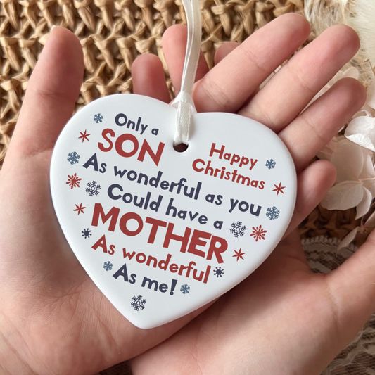 White ceramic heart-shaped Christmas ornament held in a hand as shown in 31.jpg. The ornament features red and blue snowflakes with the printed text: Only a SON As wonderful as you Could have a MOTHER As wonderful As me! Happy Christmas. It hangs from a delicate white ribbon.
