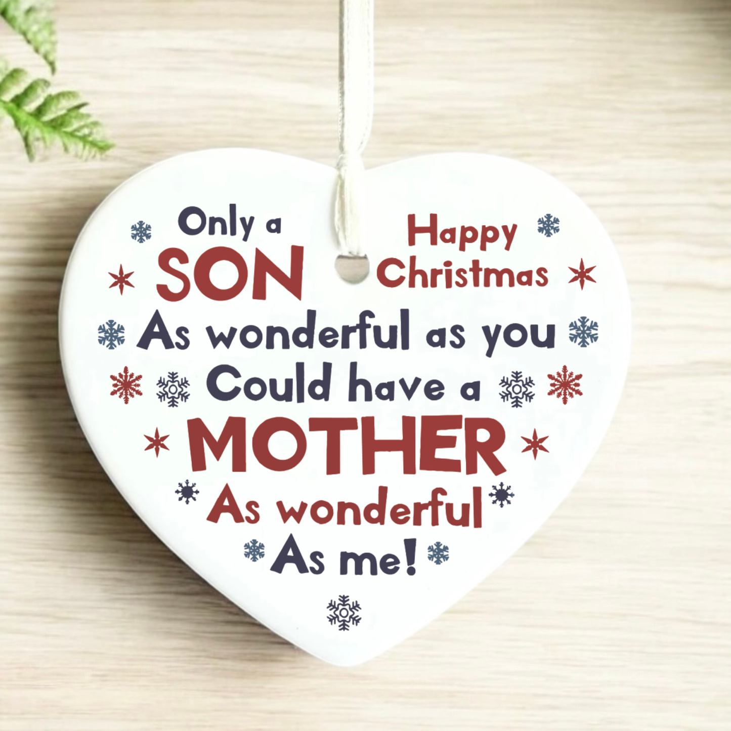 A heart-shaped white ceramic Christmas ornament hanging on a light wood surface, printed with the message 'Only a SON As wonderful as you Could have a MOTHER As me!' in red and navy lettering with small snowflake accents.