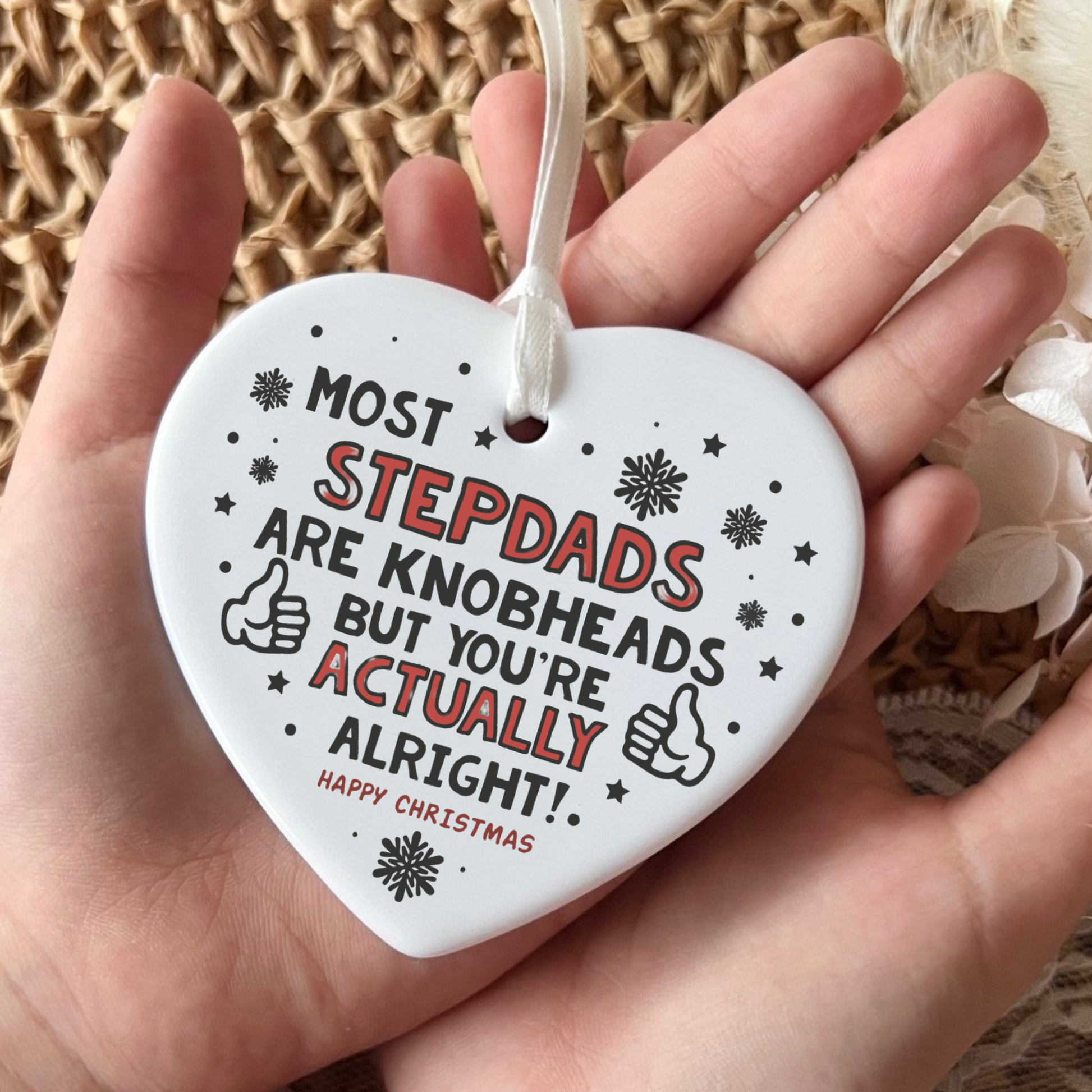A white heart-shaped ceramic Christmas ornament held in a hand, featuring the humorous text 'MOST STEPDADS ARE KNOBHEADS BUT YOU'RE ACTUALLY ALRIGHT!' in black and red lettering with snowflake and thumbs-up illustrations.