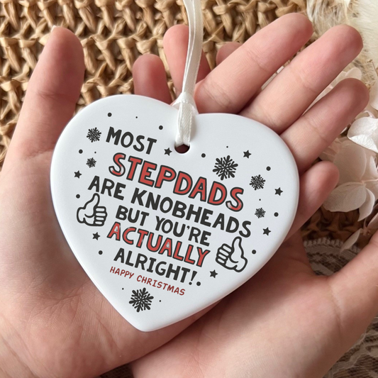 A white heart-shaped ceramic Christmas ornament held in a hand, featuring the humorous text 'MOST STEPDADS ARE KNOBHEADS BUT YOU'RE ACTUALLY ALRIGHT!' in black and red lettering with snowflake and thumbs-up illustrations.