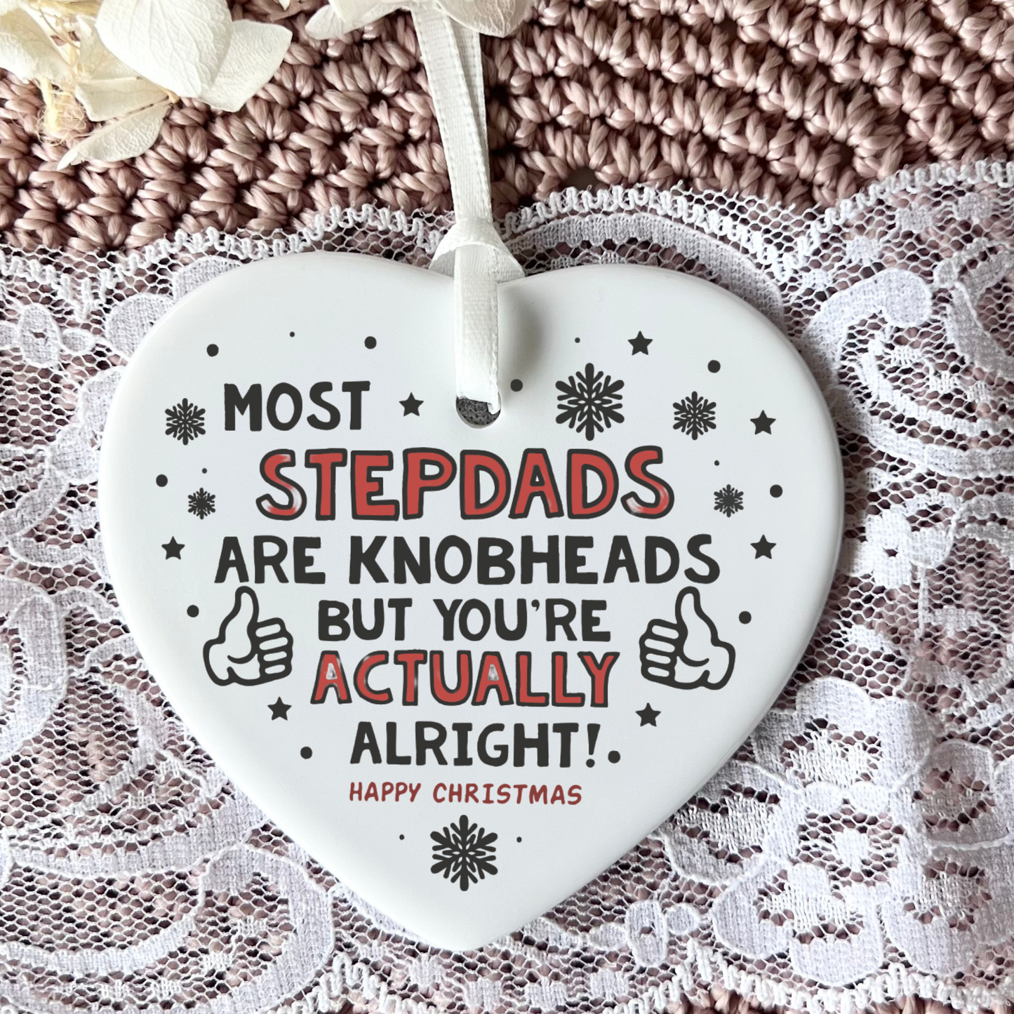 A white heart-shaped Christmas ornament with a hanging ribbon, featuring the humorous text 'MOST STEPDADS ARE KNOBHEADS BUT YOU'RE ACTUALLY ALRIGHT! HAPPY CHRISTMAS' in black and red lettering, displayed on a lace and knit background.