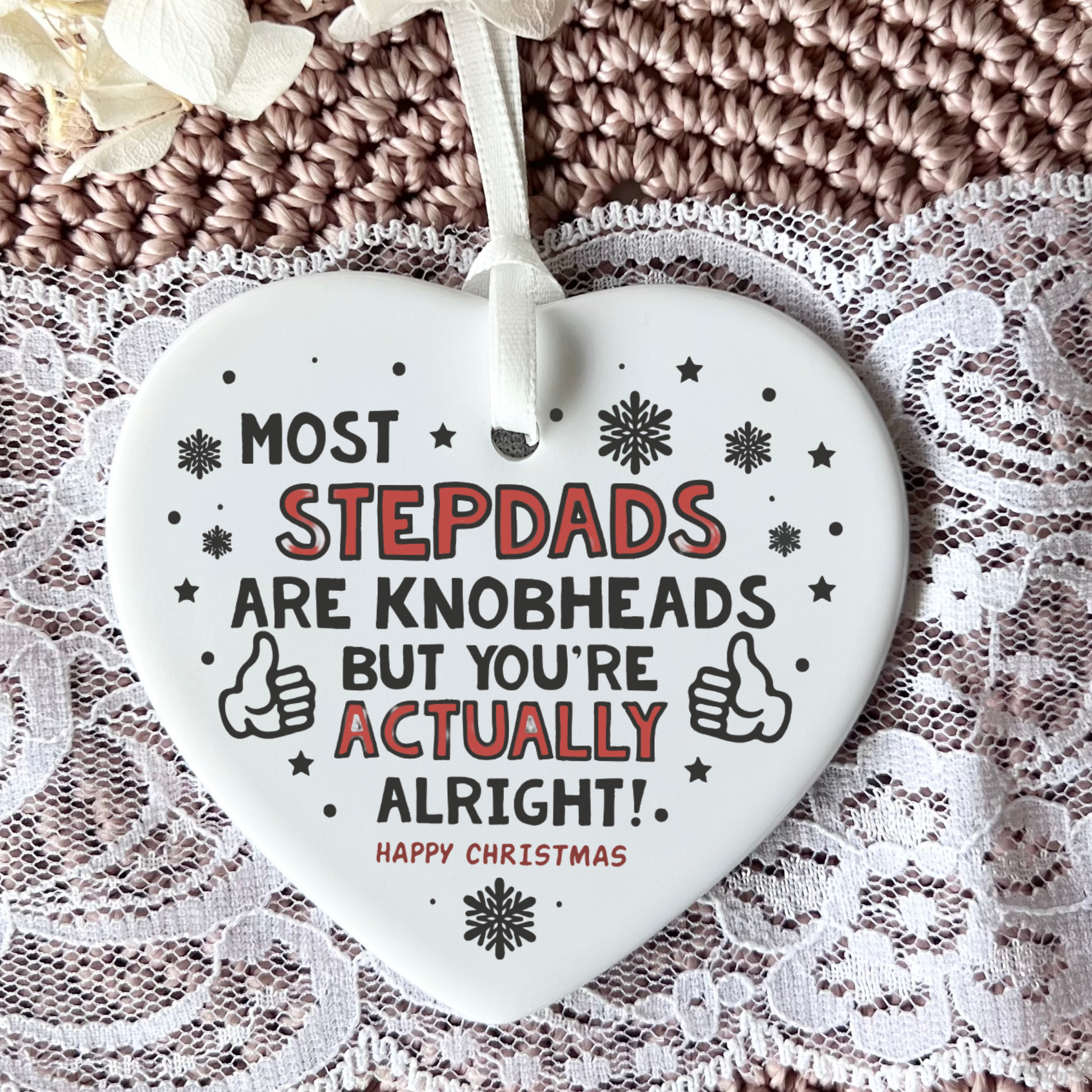 A white heart-shaped Christmas ornament with a hanging ribbon, featuring the humorous text 'MOST STEPDADS ARE KNOBHEADS BUT YOU'RE ACTUALLY ALRIGHT! HAPPY CHRISTMAS' in black and red lettering, displayed on a lace and knit background.