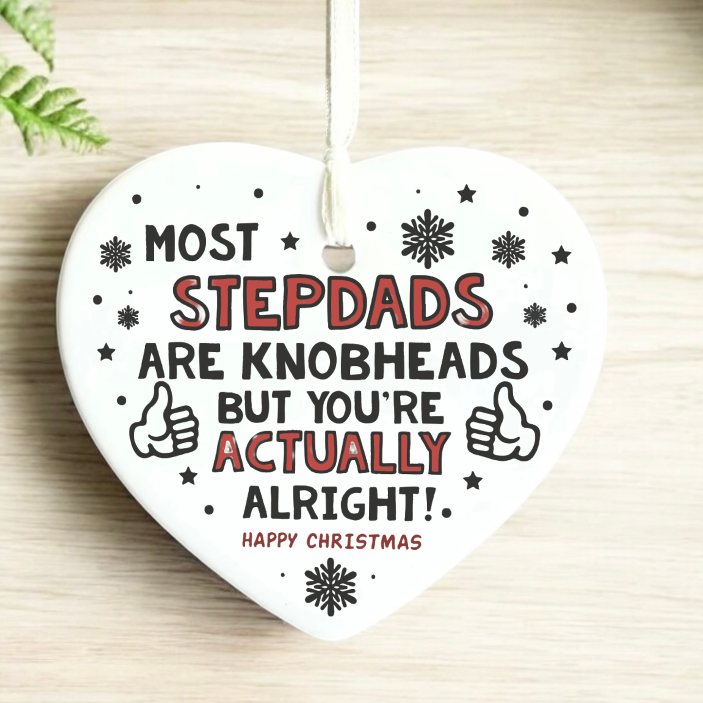 A heart-shaped white ceramic Christmas ornament with a hanging loop, featuring the humorous text 'MOST STEPDADS ARE KNOBHEADS BUT YOU'RE ACTUALLY ALRIGHT!' in black and red lettering, surrounded by black snowflakes and thumbs-up icons, displayed on a light wood surface.