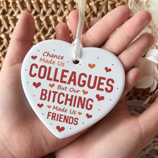 White ceramic heart-shaped Christmas ornament featuring a glossy finish. The ornament is printed with a humorous message in red and orange text that reads: "Chance Made Us COLLEAGUES But Our BITCHING Made Us FRIENDS". The design is adorned with scattered red and orange love hearts and small dots, finished with a delicate white satin hanging ribbon.