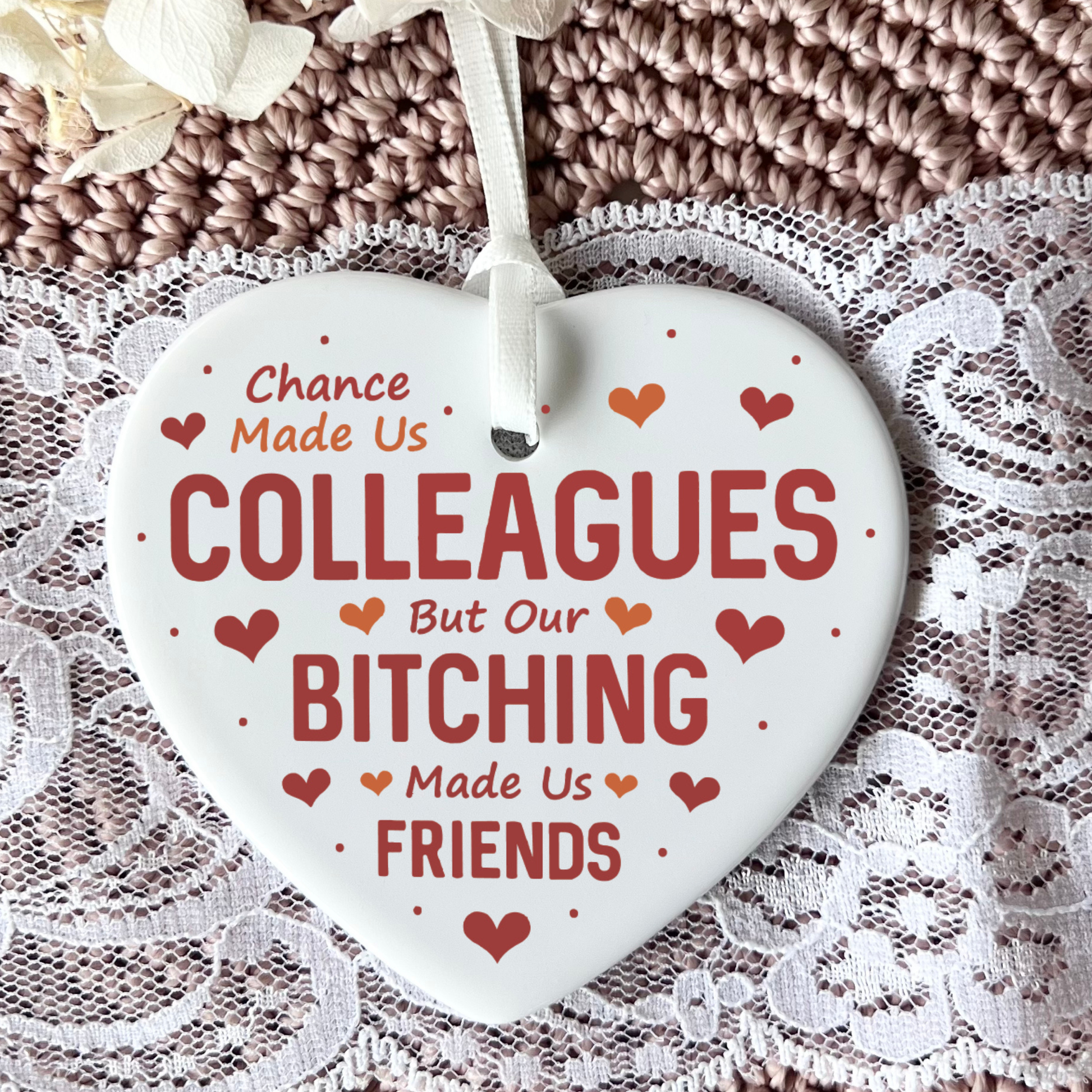 A white heart-shaped ceramic ornament with red and orange text reading 'Chance Made Us Colleagues But Our Bitching Made Us Friends,' hanging from a white ribbon against a brown knit blanket and white lace background.