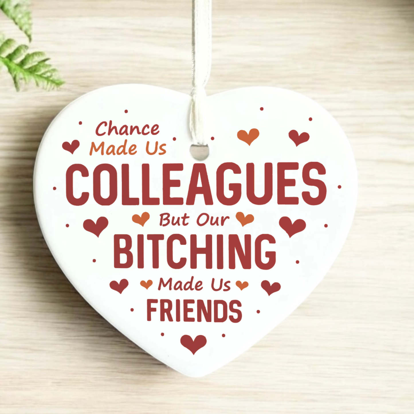 A white heart-shaped ceramic ornament hanging from a white string, printed with the humorous phrase 'Chance Made Us COLLEAGUES But Our BITCHING Made Us FRIENDS' in dark red lettering, surrounded by small red hearts and dots, on a light wood background.