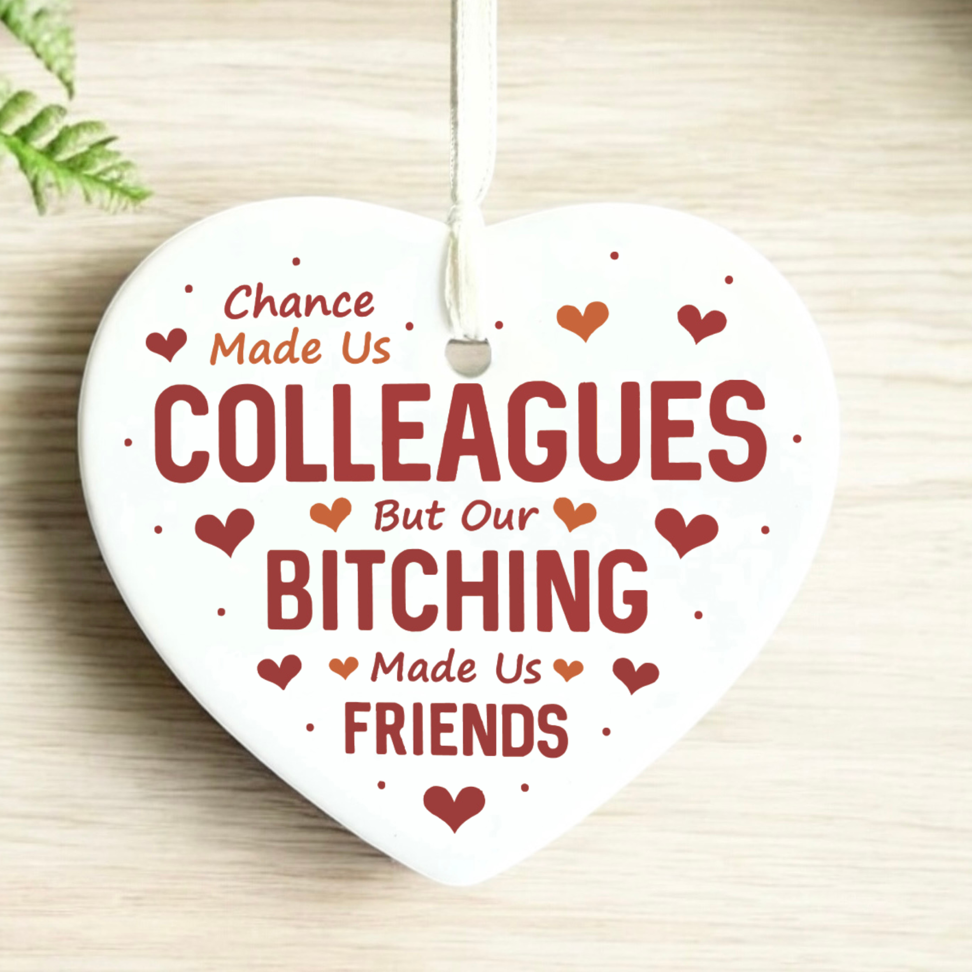 A white heart-shaped ceramic ornament hanging from a white string, printed with the humorous phrase 'Chance Made Us COLLEAGUES But Our BITCHING Made Us FRIENDS' in dark red lettering, surrounded by small red hearts and dots, on a light wood background.