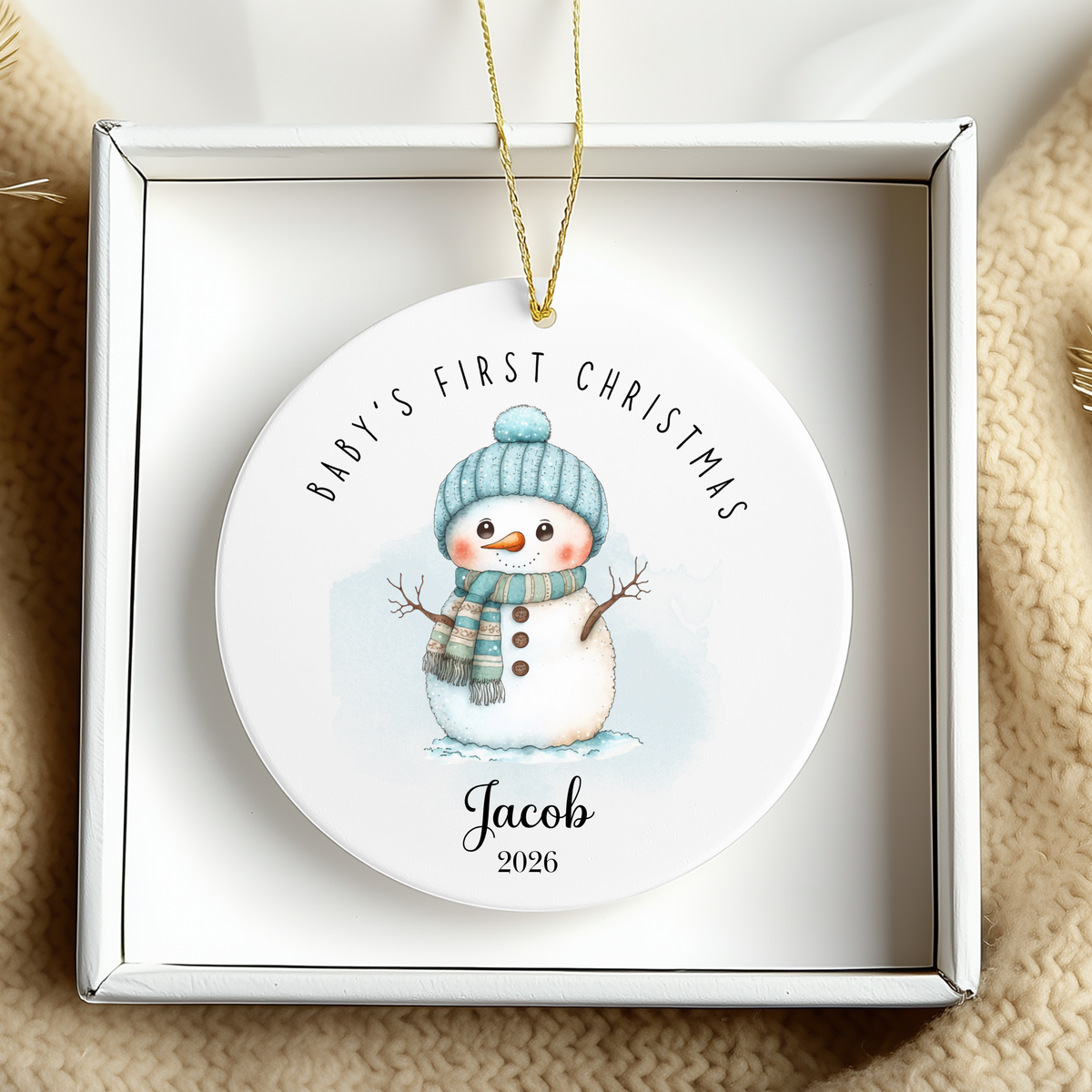 A personalized round ceramic Christmas ornament in a white gift box, featuring a watercolor snowman wearing a blue knit hat and scarf with the text 'BABY'S FIRST CHRISTMAS' and the name 'Jacob 2026'.