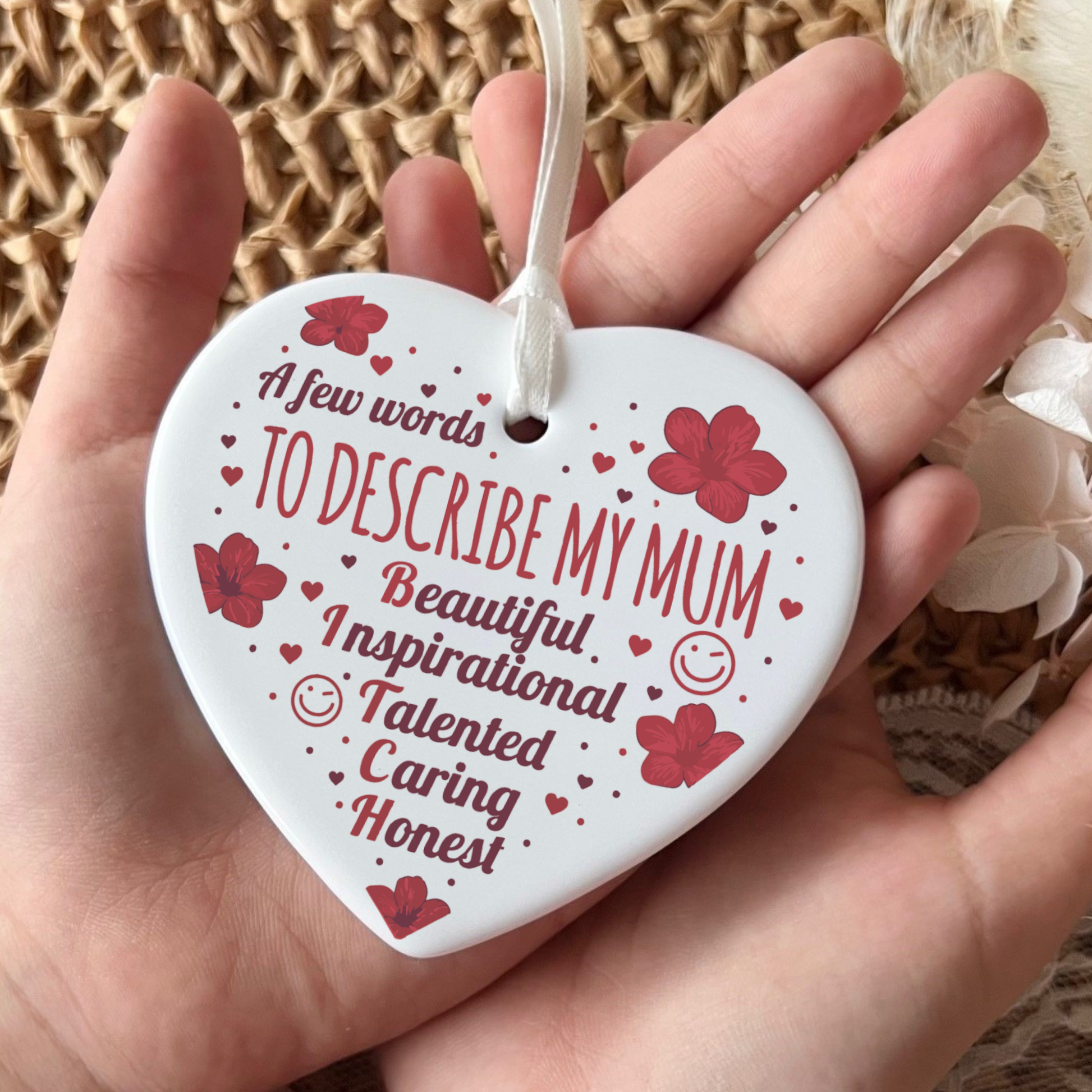 A white heart-shaped ceramic ornament with red floral and heart designs, held in a hand. The ornament features personalized text that reads 'A few words TO DESCRIBE MY MUM Beautiful Inspirational Talented Caring Honest' along with small smiley faces.