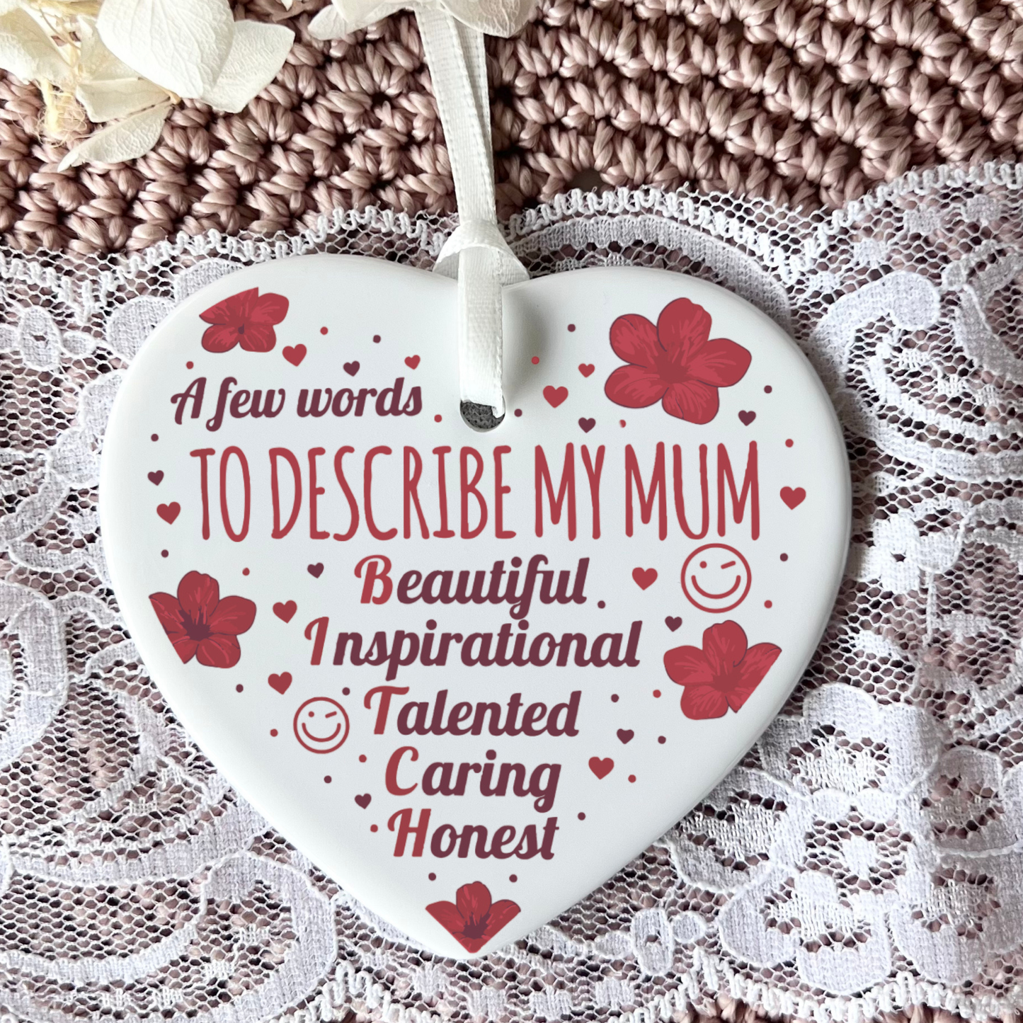 A heart-shaped white ceramic ornament with red text and floral accents, hanging from a white ribbon over a lace and knitted textile background.