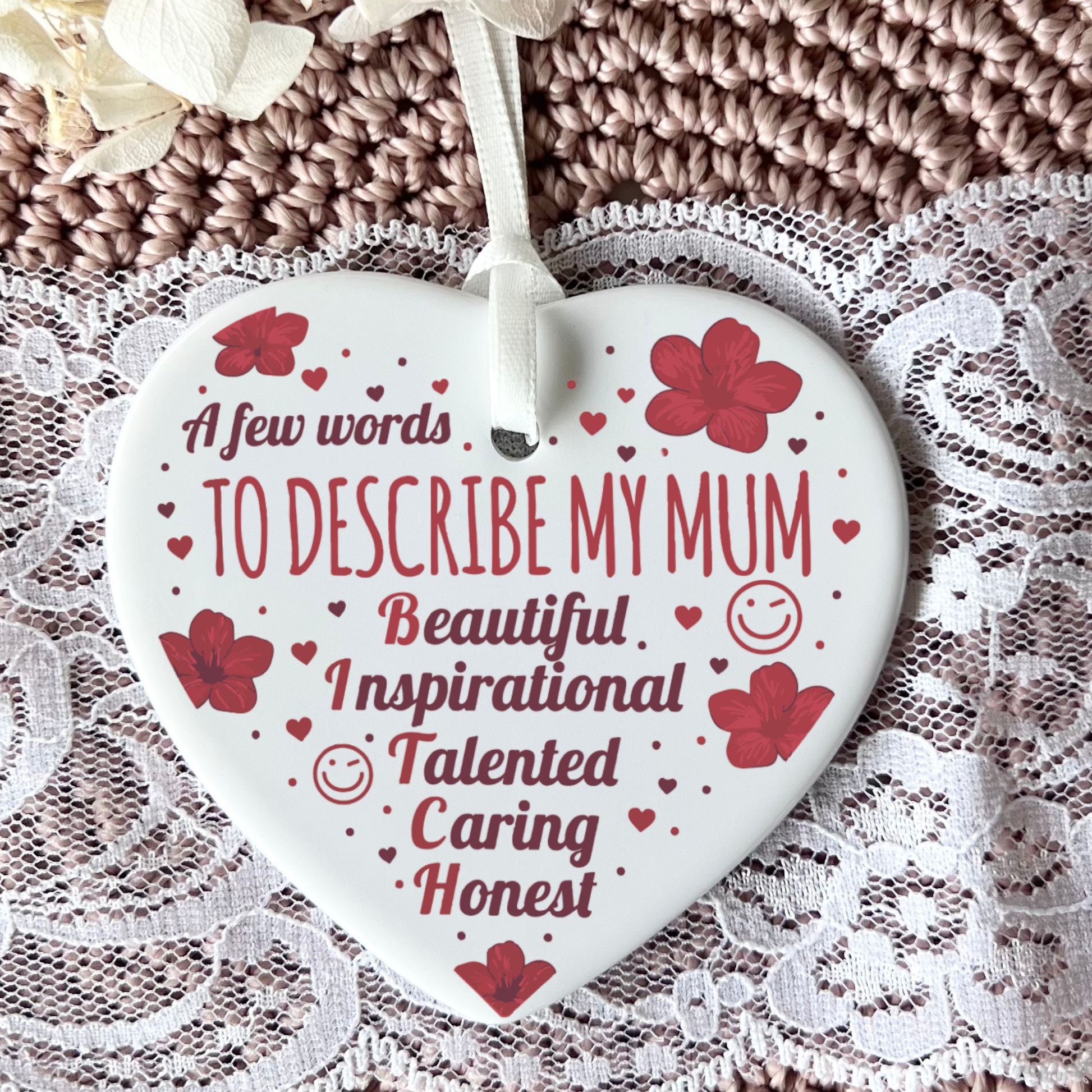 A heart-shaped white ceramic ornament with red text and floral accents, hanging from a white ribbon over a lace and knitted textile background.