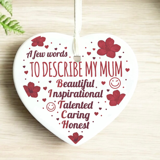 A heart-shaped white ceramic hanging ornament with red floral designs and the printed message 'TO DESCRIBE MY MUM Beautiful Inspirational Talented Caring Honest' on a light wood surface.