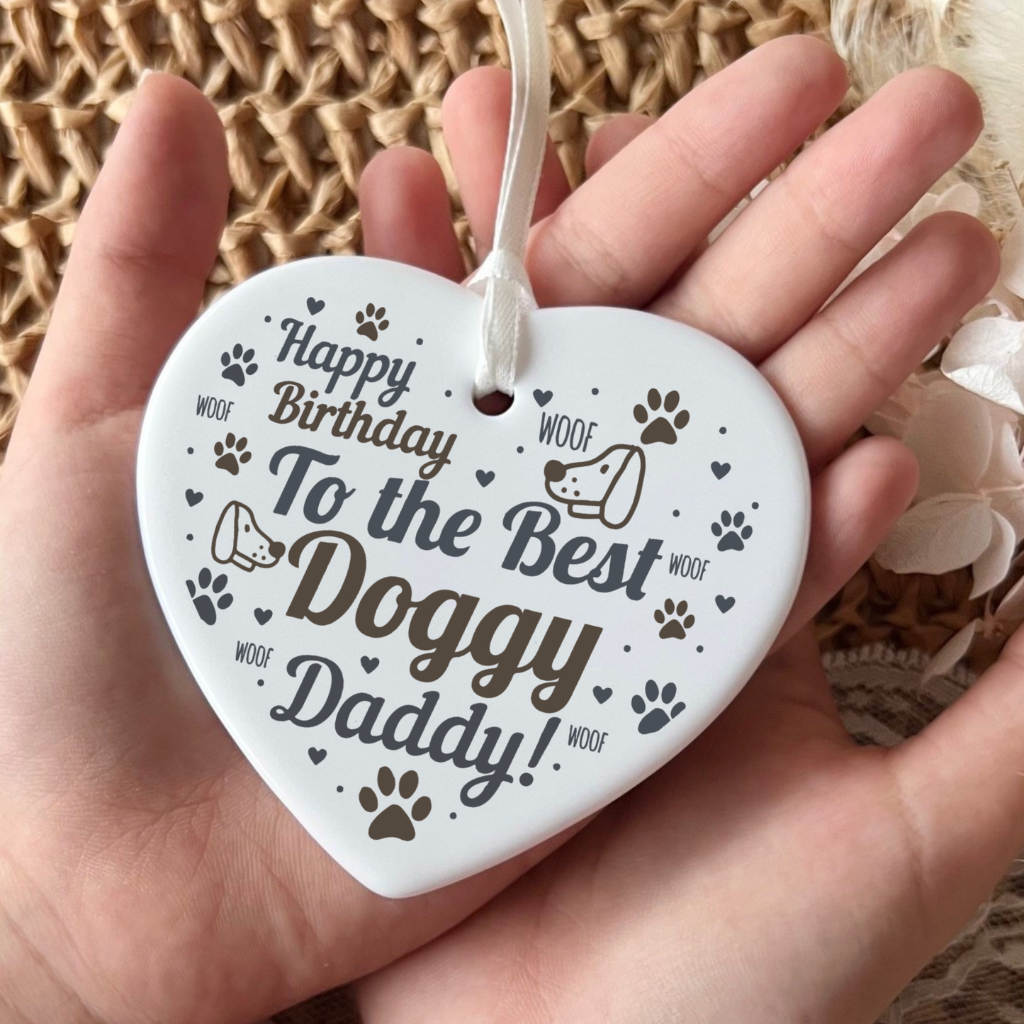 A white heart-shaped ceramic dog birthday ornament held in a person's hand, printed with the text 'Happy Birthday To the Best Doggy Daddy!' and small dog paw prints and doodles.