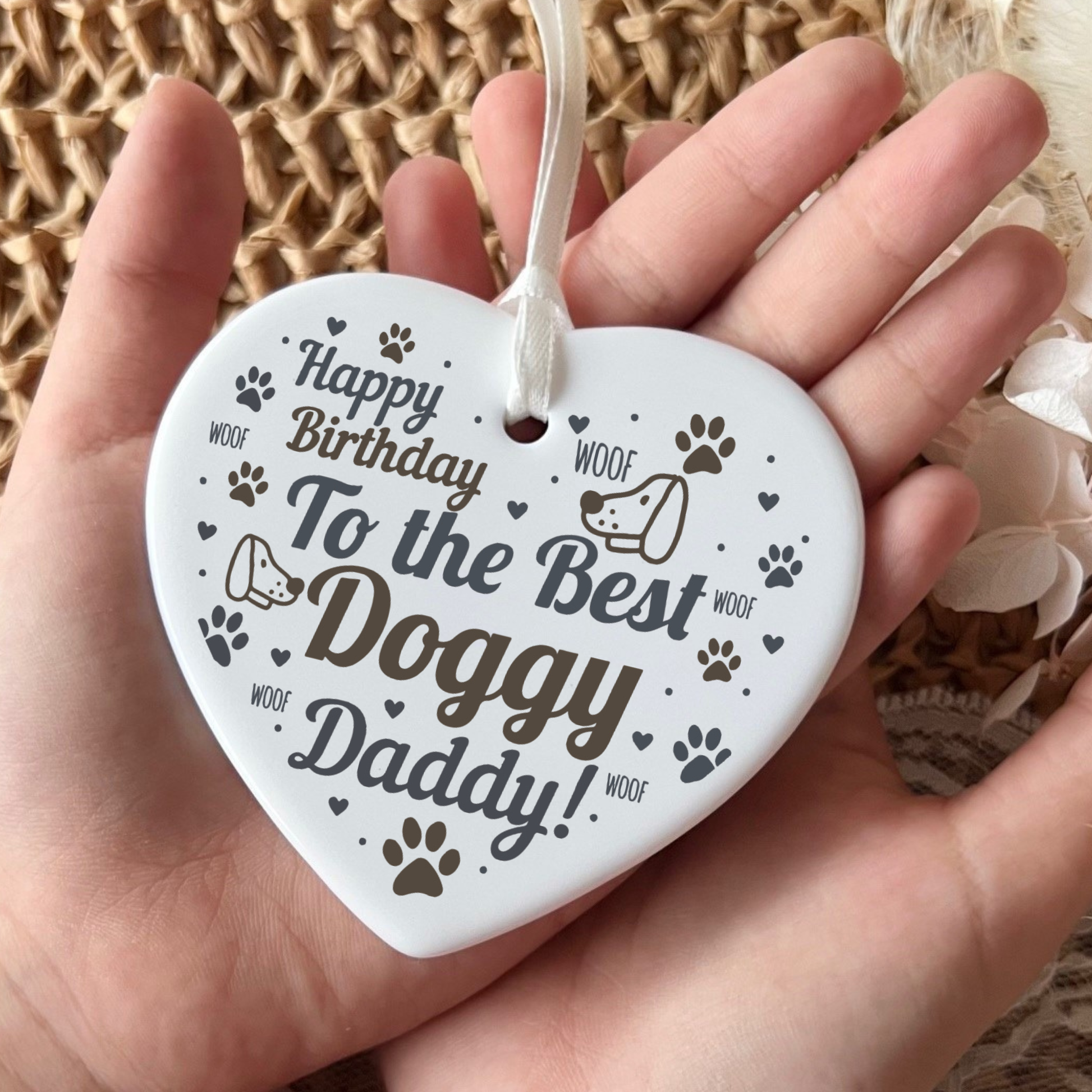A white heart-shaped ceramic dog birthday ornament held in a person's hand, printed with the text 'Happy Birthday To the Best Doggy Daddy!' and small dog paw prints and doodles.