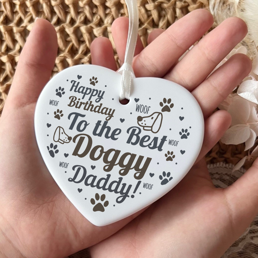 A white heart-shaped ceramic dog birthday ornament held in a person's hand, printed with the text 'Happy Birthday To the Best Doggy Daddy!' and small dog paw prints and doodles.
