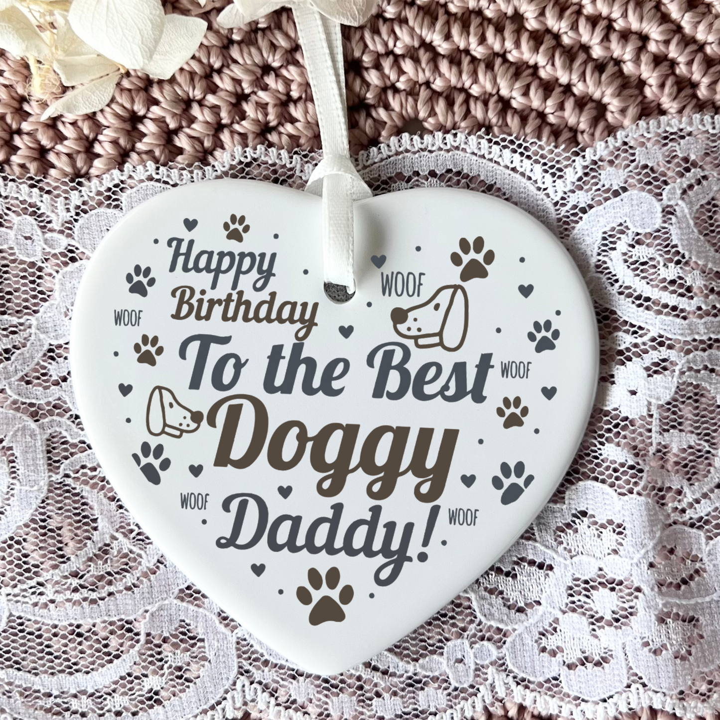 A heart-shaped white ceramic dog memorial plaque hanging from a white ribbon, featuring the text 'Happy Birthday To the Best Doggy Daddy!' in dark brown lettering, surrounded by paw prints, hearts, and dog illustrations. The plaque is displayed on a white lace surface with a brown knitted blanket in the background.