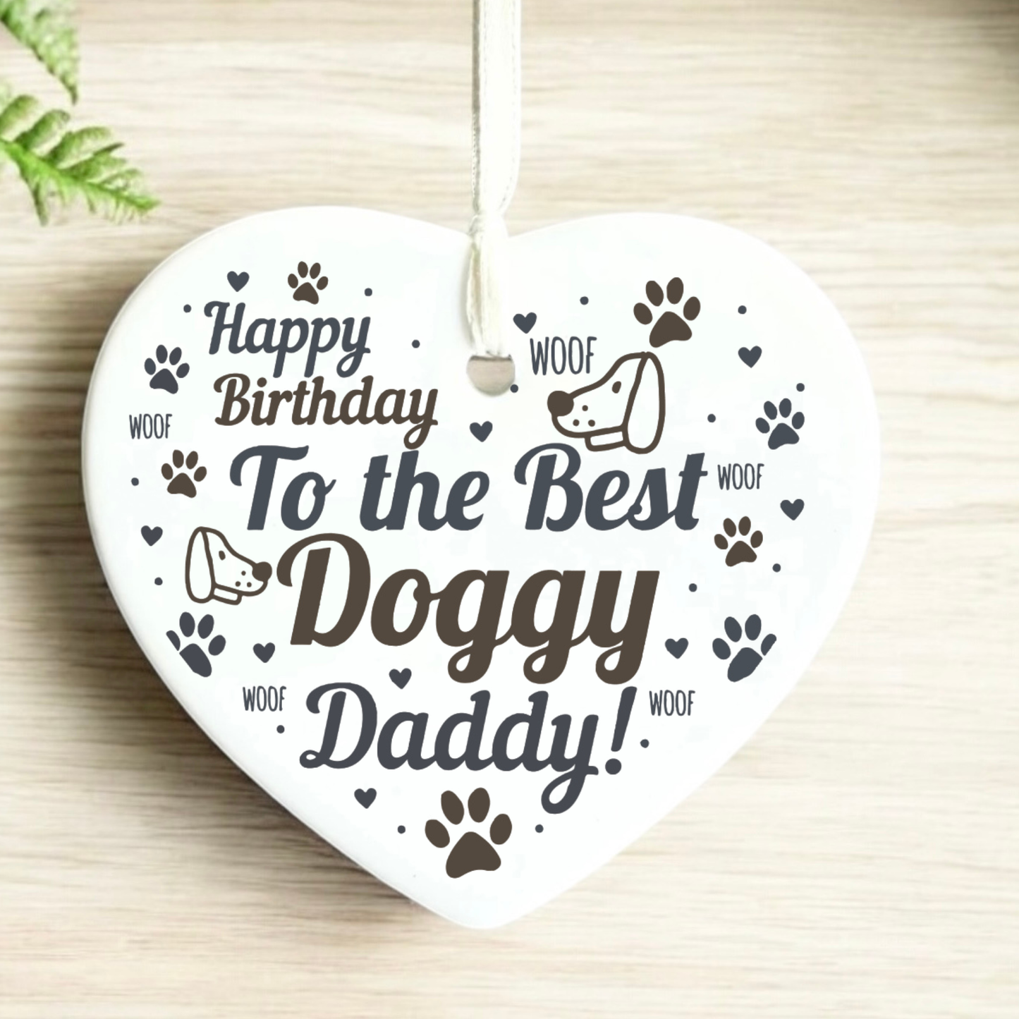 A heart-shaped white ceramic dog memorial ornament hanging from a white ribbon, printed with 'Happy Birthday To the Best Doggy Daddy!' in dark brown lettering, accompanied by paw prints, hearts, and cartoon dog faces on a light wood background.
