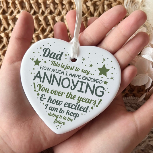A white heart-shaped ceramic ornament held in a hand, featuring a humorous quote for Dad in dark green text and star accents, with a white hanging ribbon.