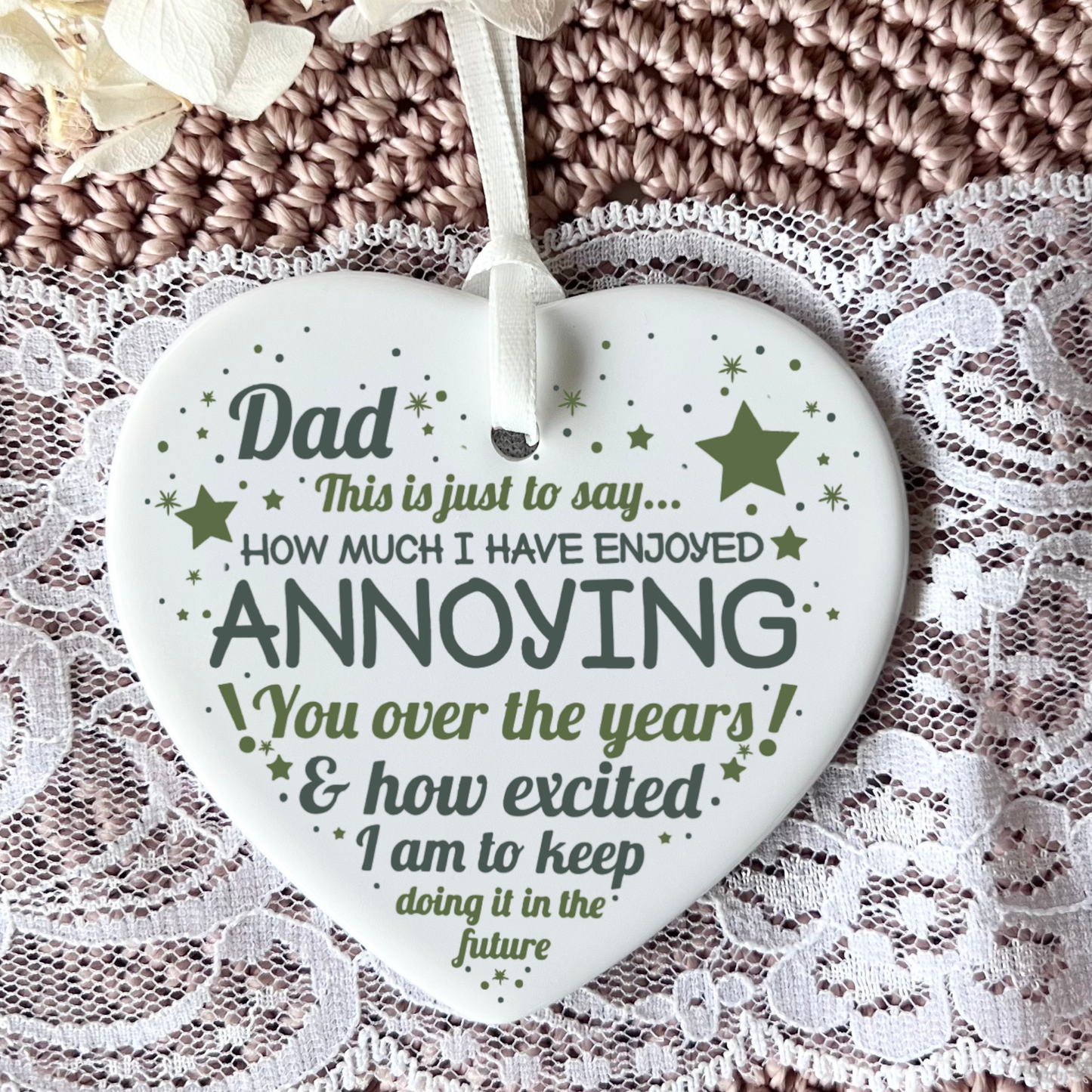 A heart-shaped white ceramic ornament with a hanging ribbon, featuring a humorous quote for Dad in dark green and gray lettering with small star accents, displayed on a textured brown knit blanket and white lace fabric.