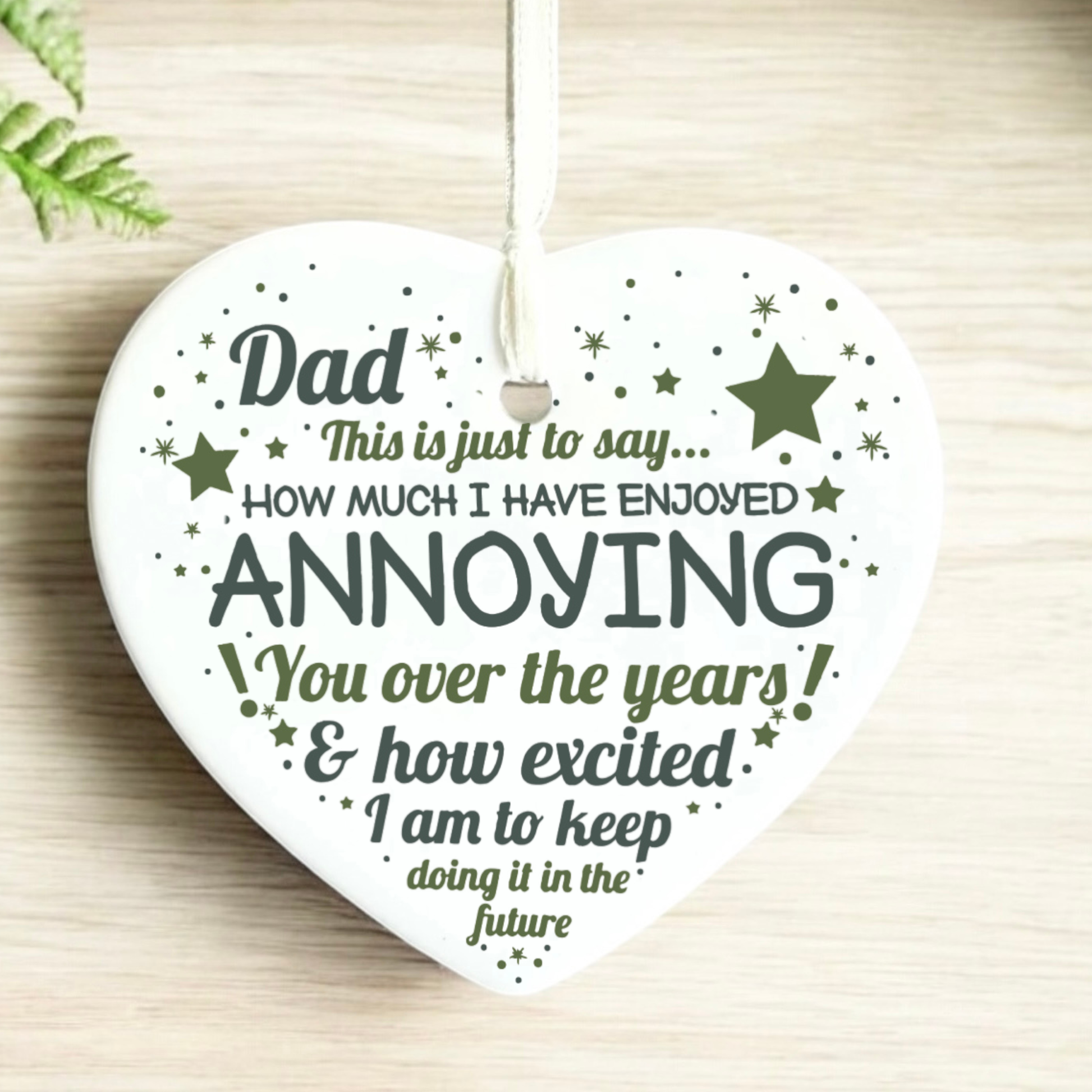 White ceramic heart-shaped Christmas ornament featuring a glossy finish. The ornament is printed with a humorous message in olive green text that reads: "Dad This is just to say... HOW MUCH I HAVE ENJOYED ANNOYING You over the years! & how excited I am to keep doing it in the future". The design is decorated with scattered olive green stars, sparkles, and dots, finished with a delicate white satin hanging ribbon.