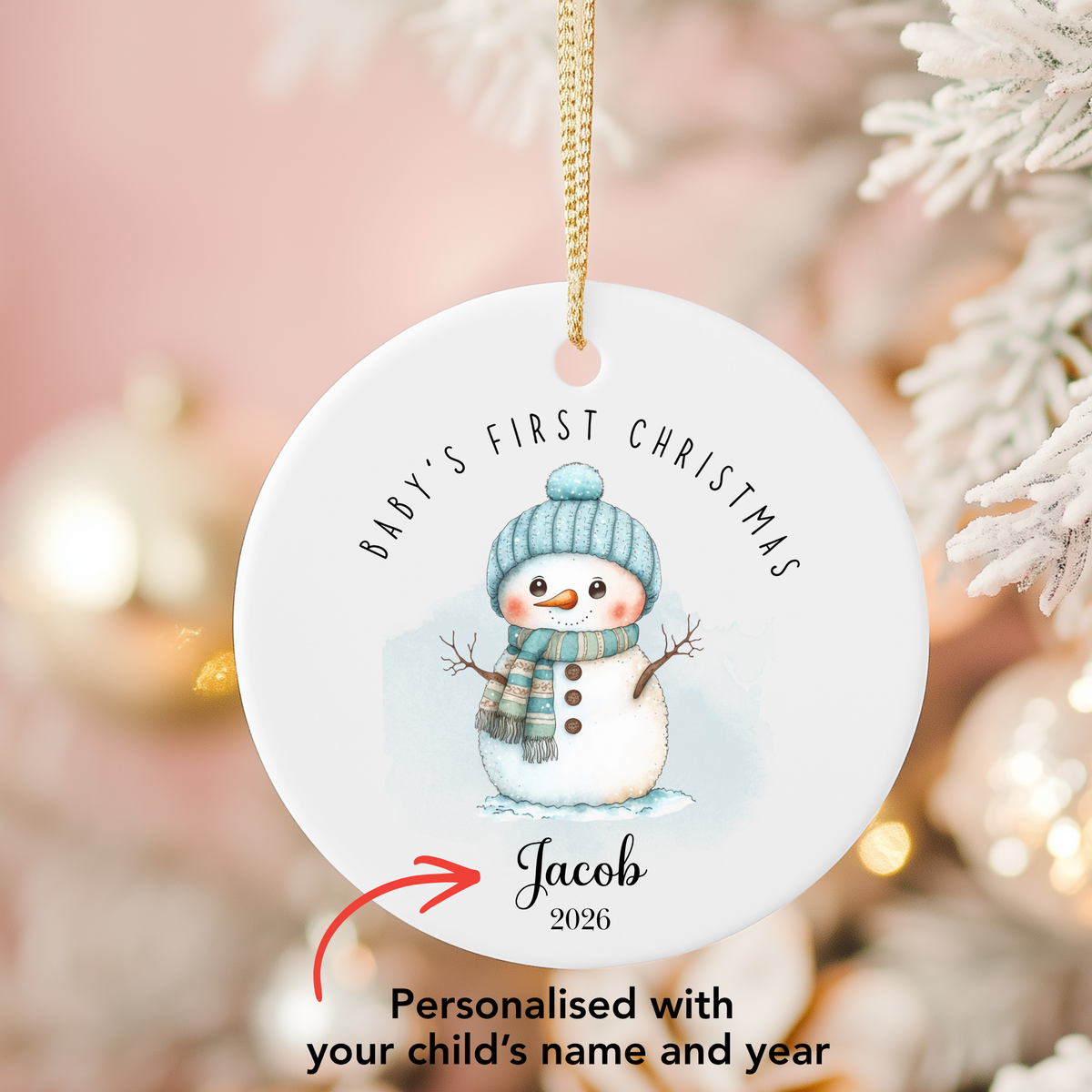 Personalized round white Christmas ornament featuring a watercolor snowman illustration with the text 'Baby's First Christmas' and a custom name 'Jacob 2026', shown hanging against a soft blurred festive background with white tree branches.