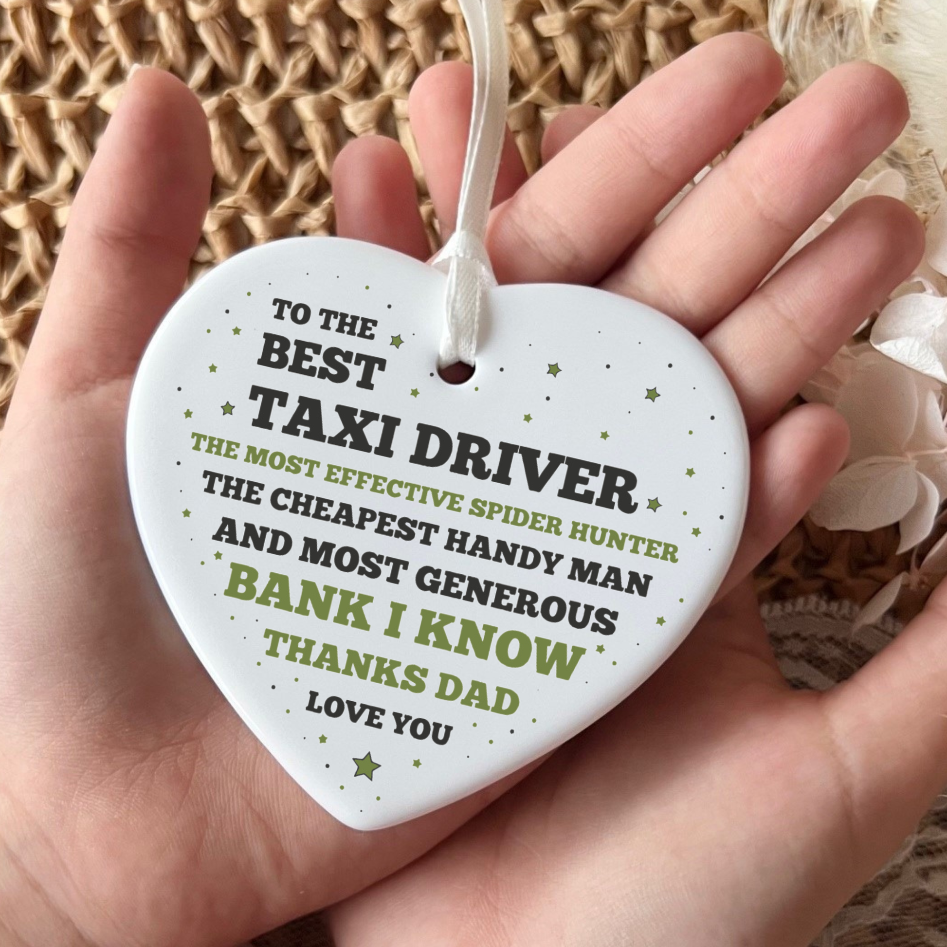 A hand holding a white ceramic heart-shaped ornament featuring a funny thank you message to a dad, with text highlighting his roles as a taxi driver, spider hunter, handyman, and bank, finished with a white ribbon.