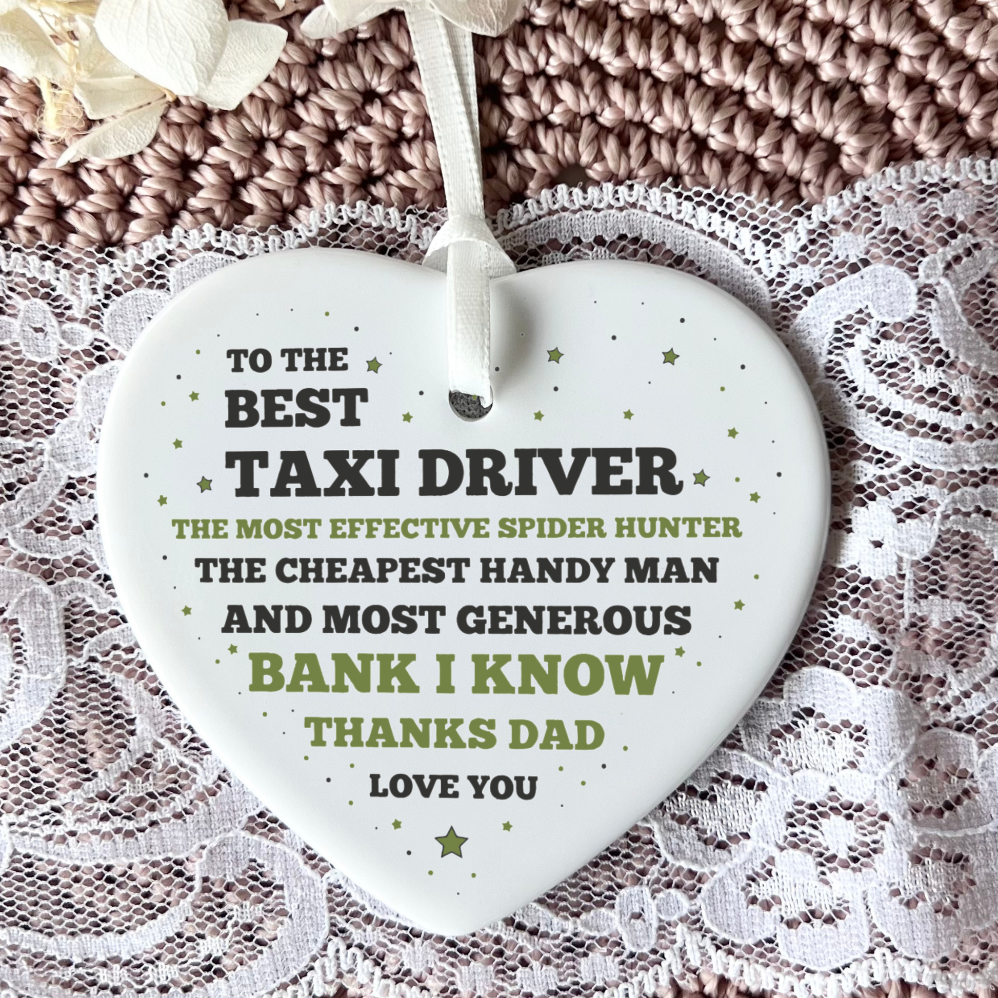 A hand holding a white ceramic heart-shaped ornament featuring a funny thank you message to a dad, with text highlighting his roles as a taxi driver, spider hunter, handyman, and bank, finished with a white ribbon.