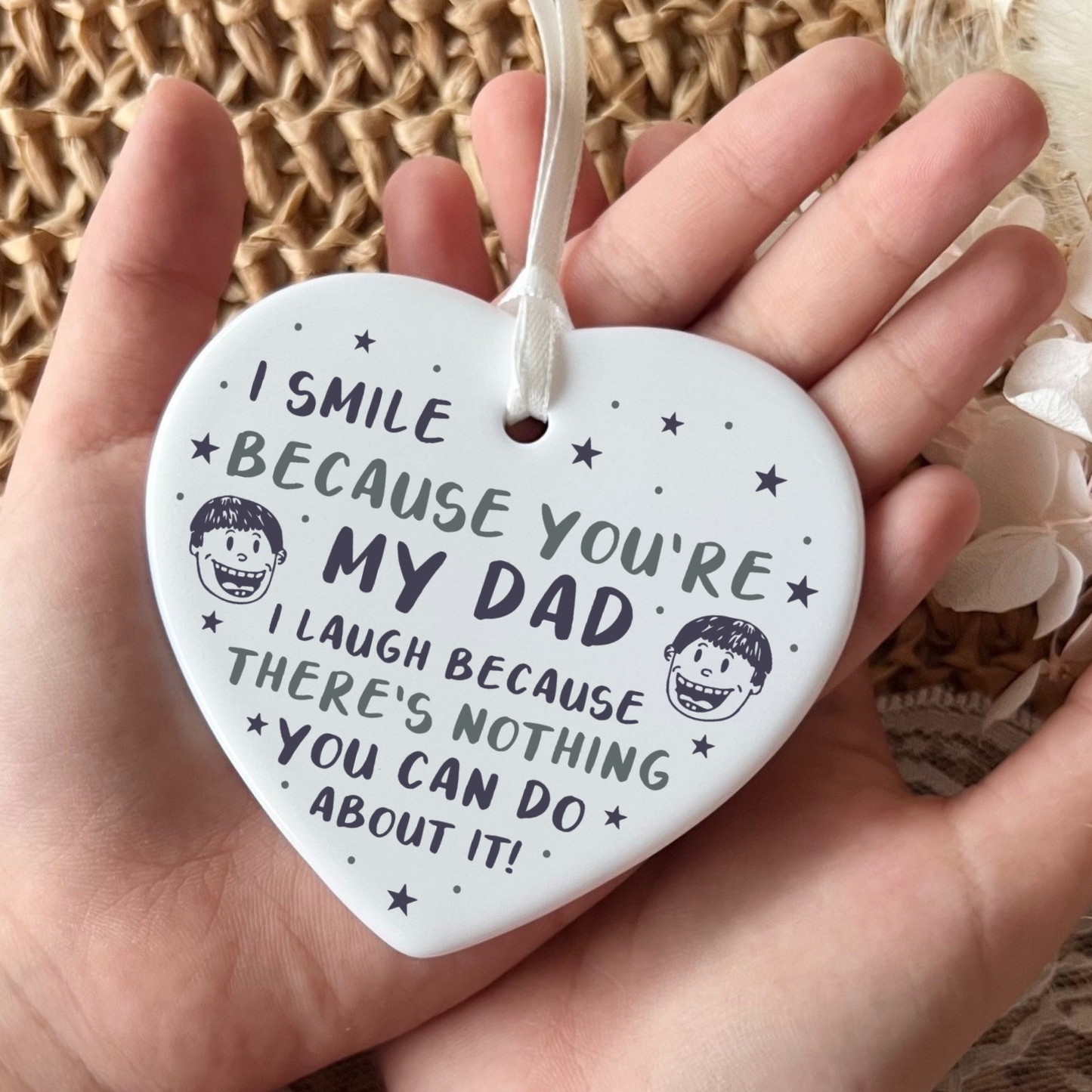 A close-up shot of a white ceramic heart-shaped hanging ornament held in two hands. The ornament displays a funny message that reads, "I smile because you're my dad. I laugh because there's nothing you can do about it!", decorated with stars and small illustrated cartoon faces.