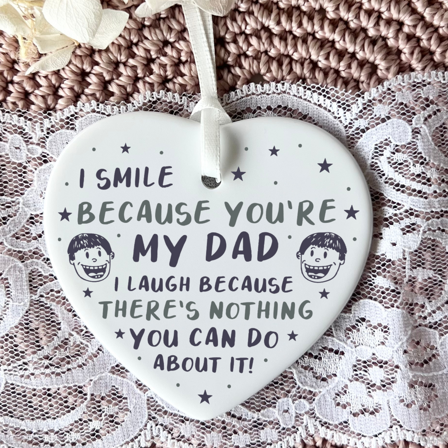A close-up shot of a white ceramic heart-shaped hanging ornament held in two hands. The ornament displays a funny message that reads, "I smile because you're my dad. I laugh because there's nothing you can do about it!", decorated with stars and small illustrated cartoon faces.