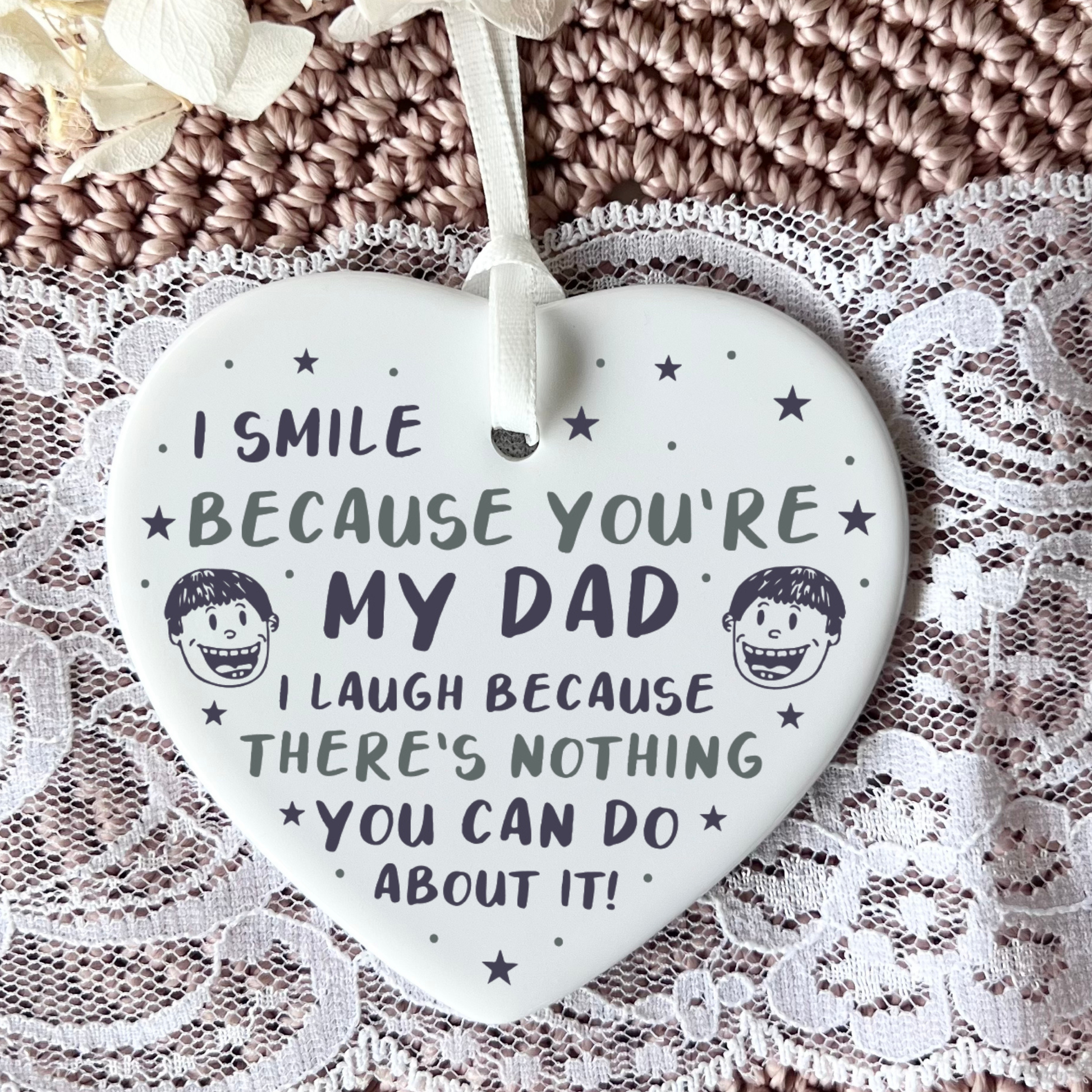 A close-up shot of a white ceramic heart-shaped hanging ornament held in two hands. The ornament displays a funny message that reads, "I smile because you're my dad. I laugh because there's nothing you can do about it!", decorated with stars and small illustrated cartoon faces.